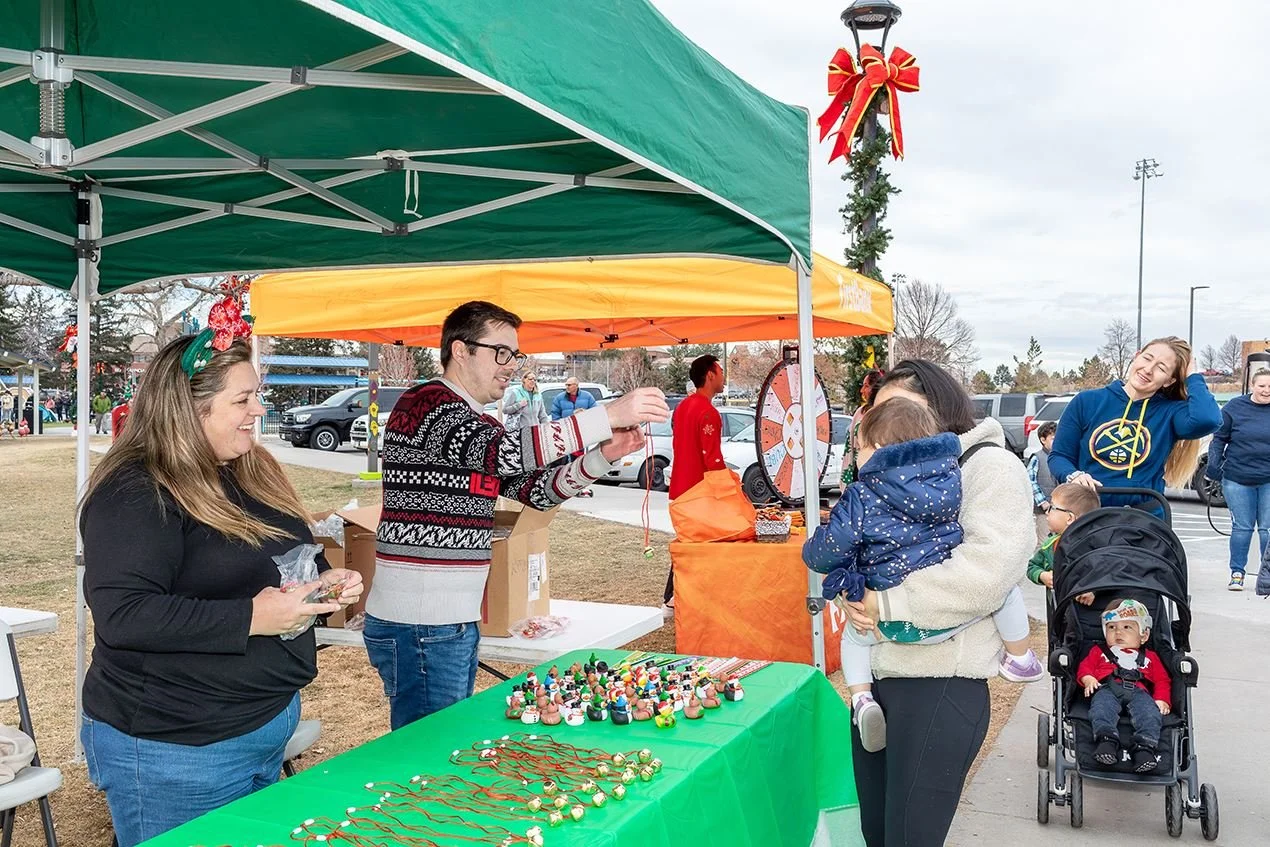 People at a Christmas-themed outdoor event with a stall selling holiday ornaments, a Christmas wreath, and a spinning wheel for prizes. One woman is holding a child, and others are smiling and browsing.