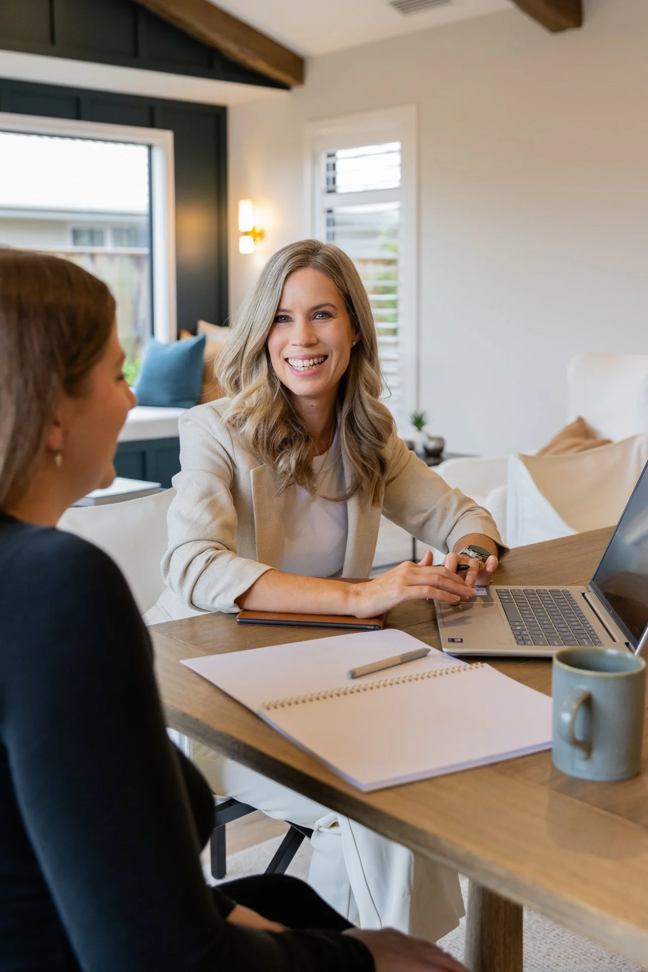 Dawn and a client having a conversation at a table in a bright, modern room. Dawn is smiling and wearing a light-colored blazer. The other woman is partially visible, wearing black. There is a laptop, a notebook, a pen, and a mug on the table.