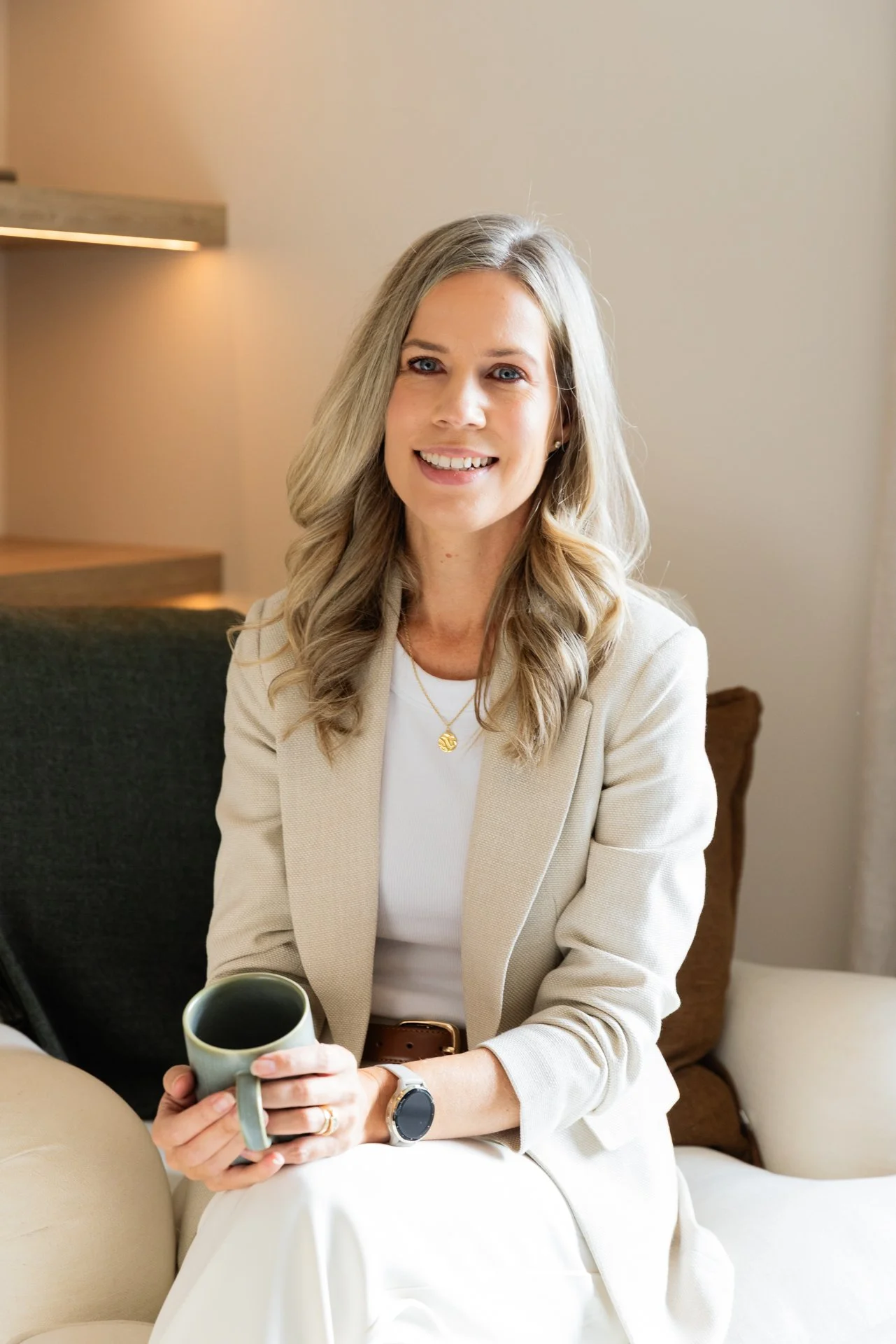 This is Dawn Toughey. She has light brown hair in loose waves, smiling, sitting on a cream-colored sofa holding a mug. She wears a beige blazer, white top, and a gold necklace with a round pendant.