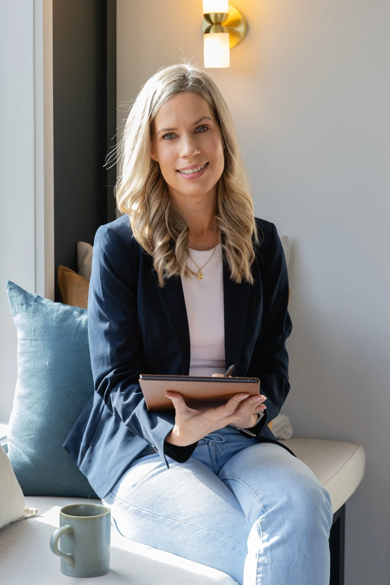 Dawn with blonde, wavy hair, wearing a white top and a navy blazer, sitting on a sofa with a blue pillow, holding a tablet, in a modern interior with a wall sconce light in the background.
