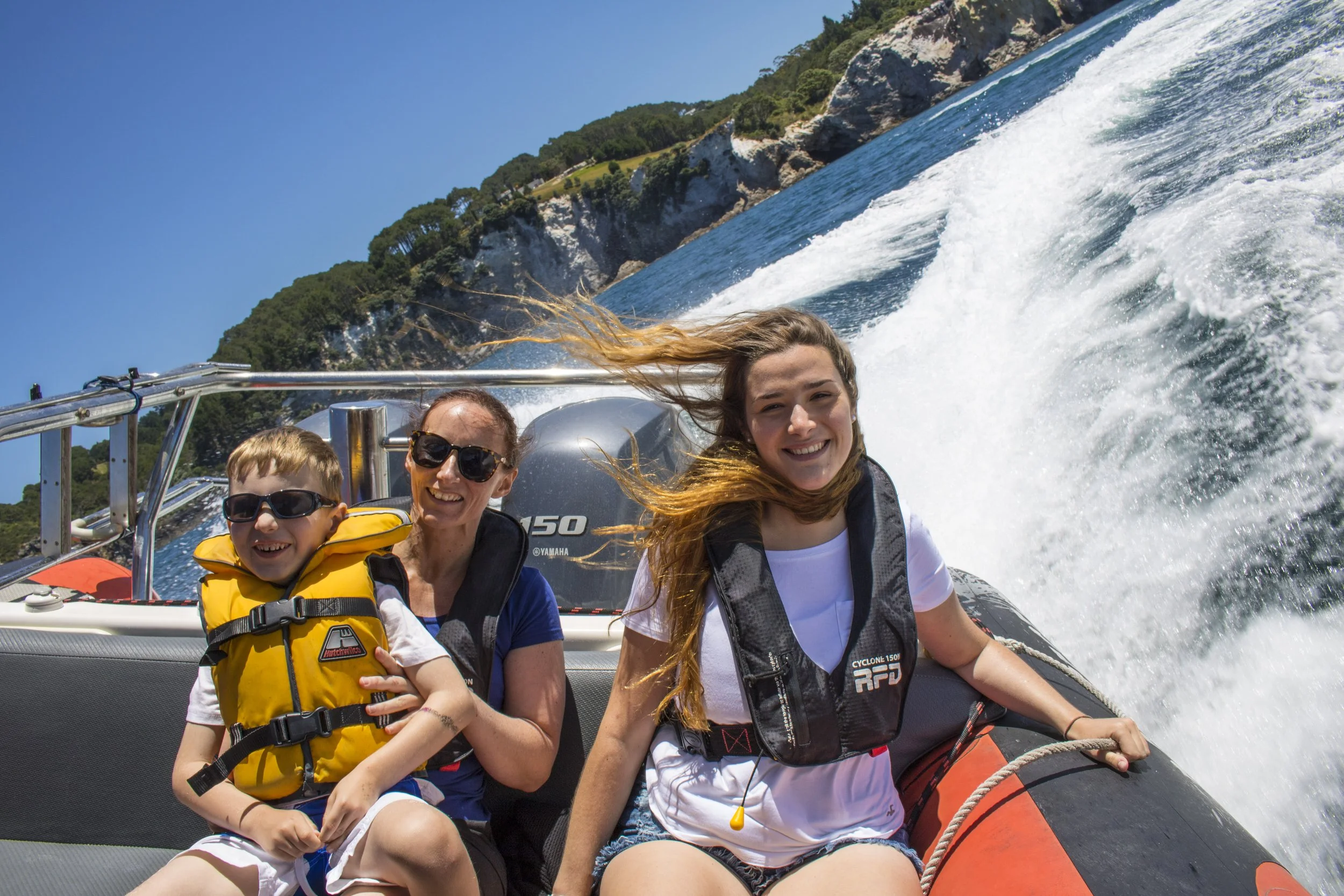 A family of three enjoying a boat ride on the water with a scenic coastal background, smiling and wearing life jackets.