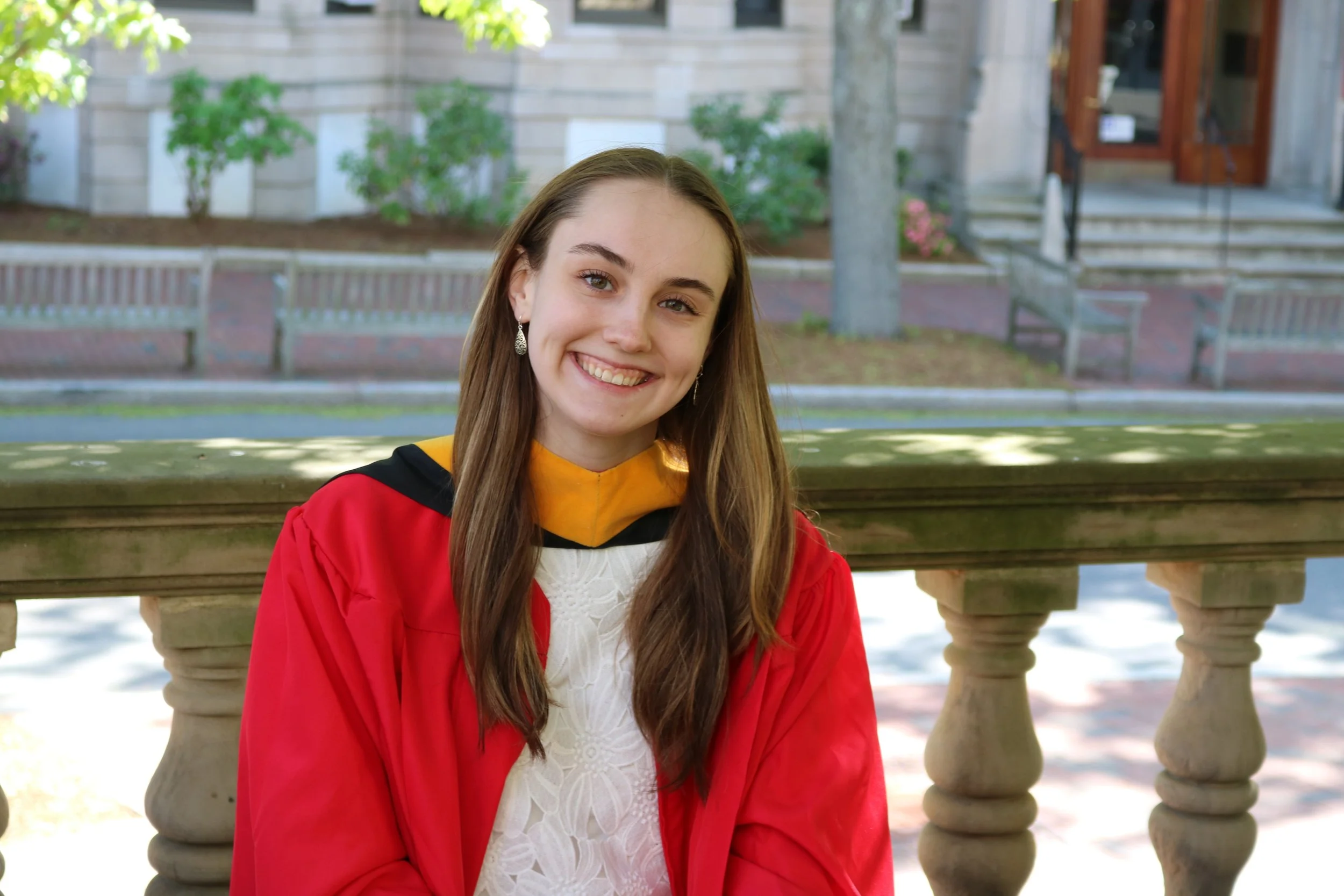 A female smiles, adorned with a graduation gown in a city garden.