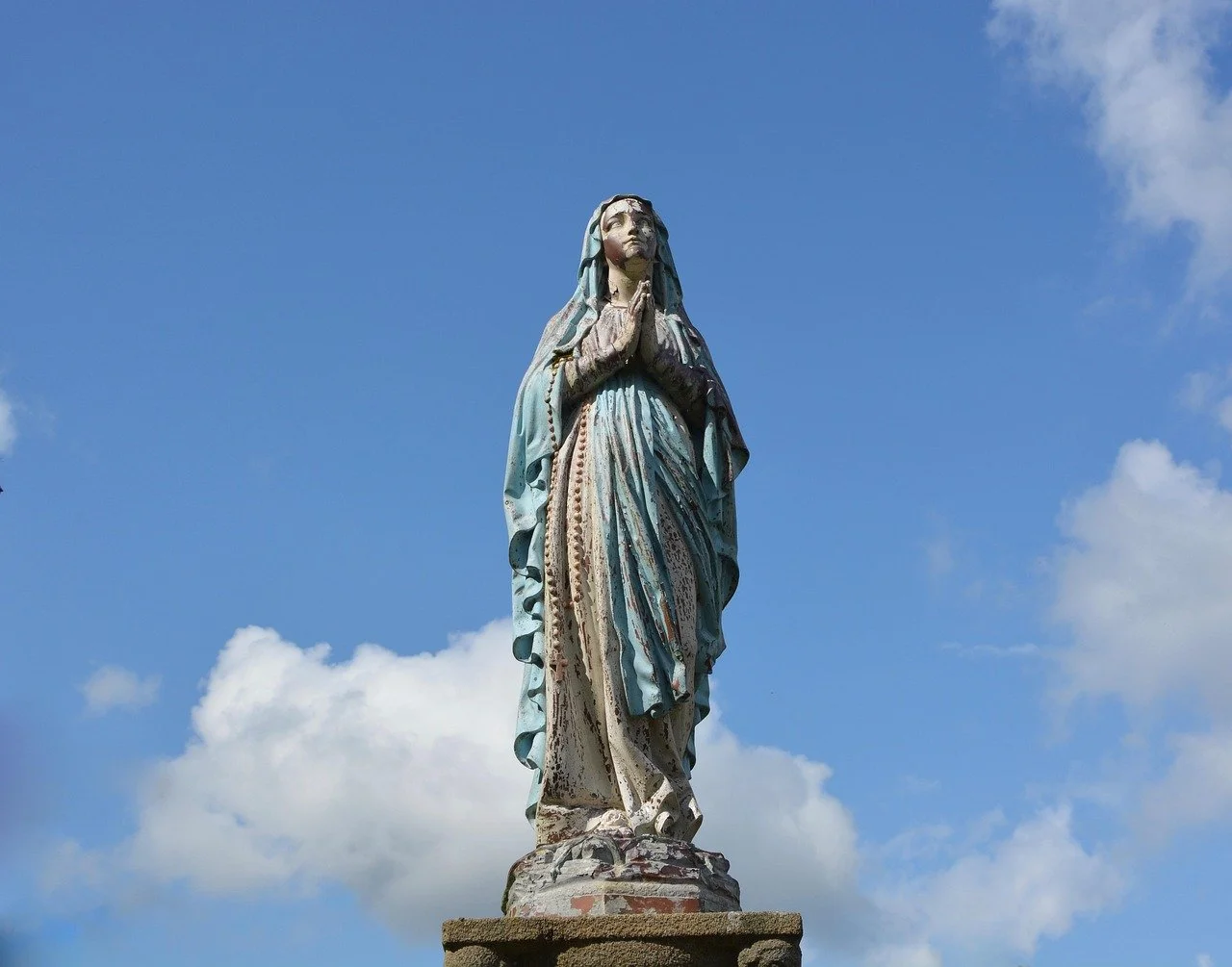 Statue of the Blessed Virgin Mary in prayer against a blue sky with clouds.