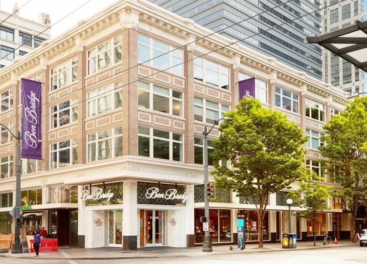 Street view of a modern multi-story building with large windows and trees outside, featuring a storefront with the sign "Ben Bridge" on the corner.