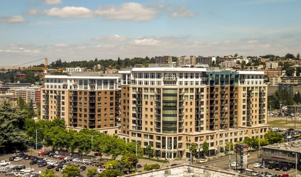 High-rise apartment building in an urban area with a parking lot and trees in the foreground, and a cityscape with construction crane and hills in the background.