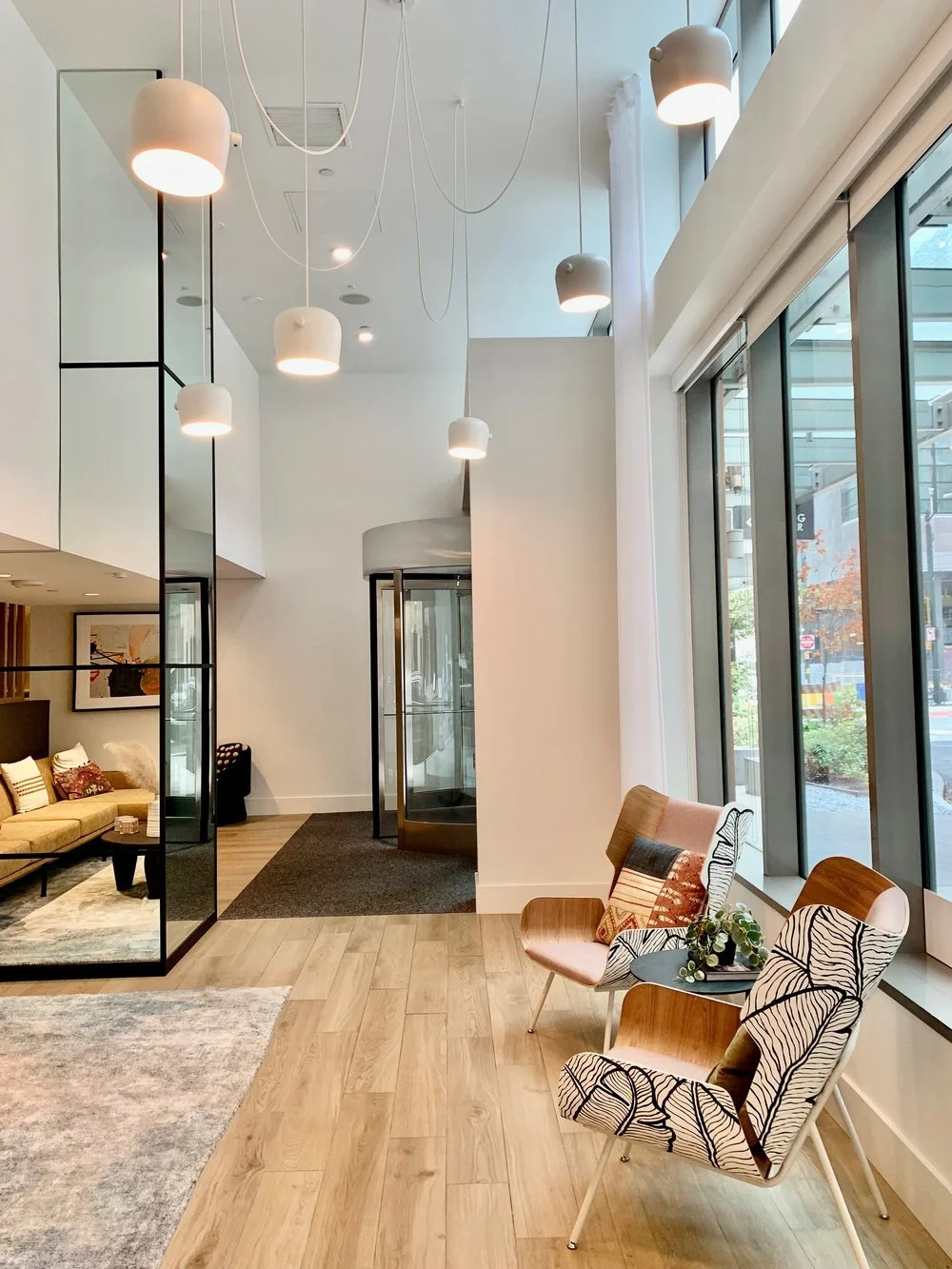 Modern hotel lobby with white walls, large windows, and wooden flooring. Features hanging white pendant lights, a beige sofa with decorative pillows, two patterned chairs by the window with a small table holding plants, and a glass revolving door in the background.