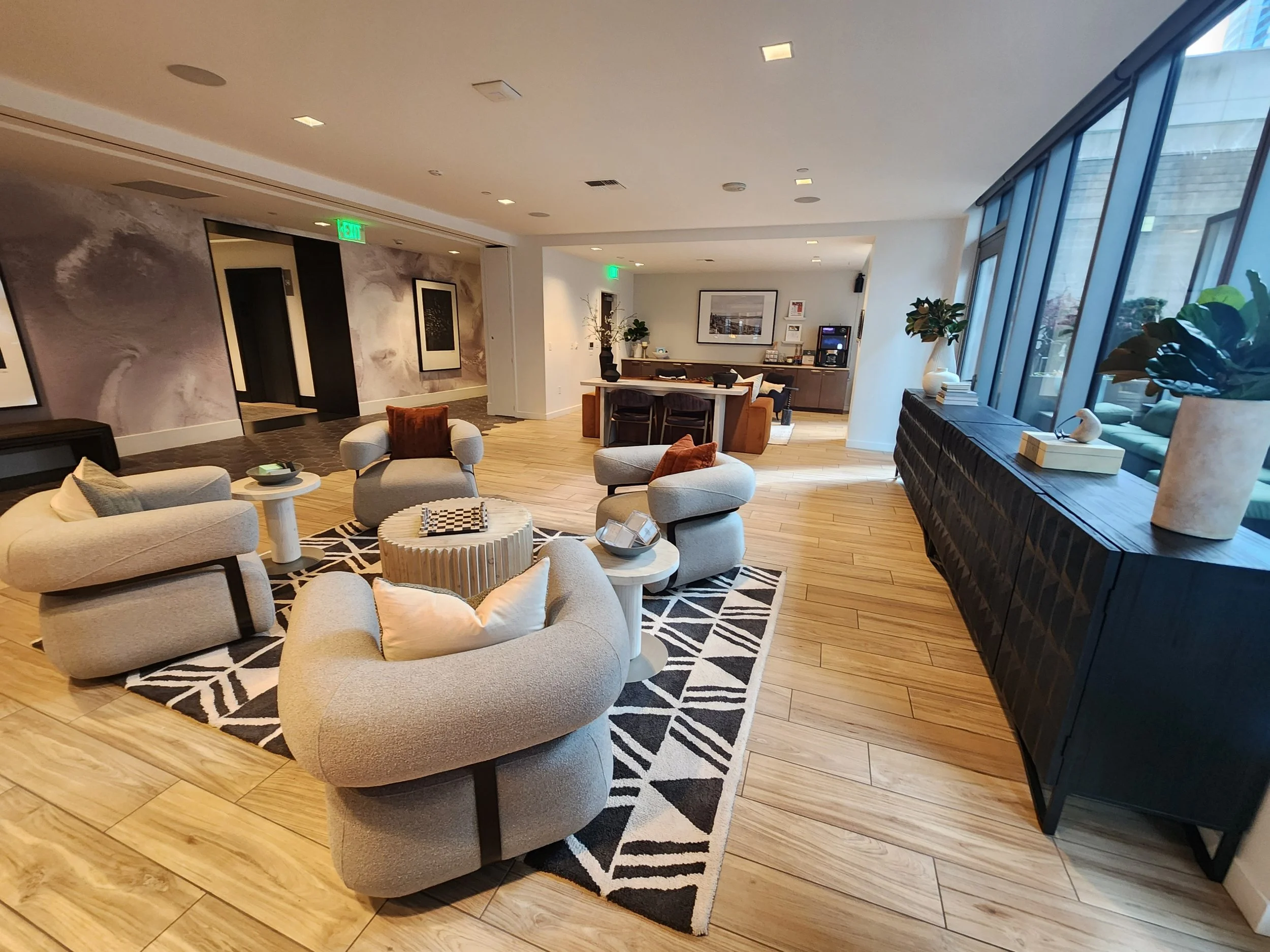 Modern hotel lobby with gray armchairs, a patterned rug, wooden floors, and large windows with decorative plants and sculptures on the windowsill.