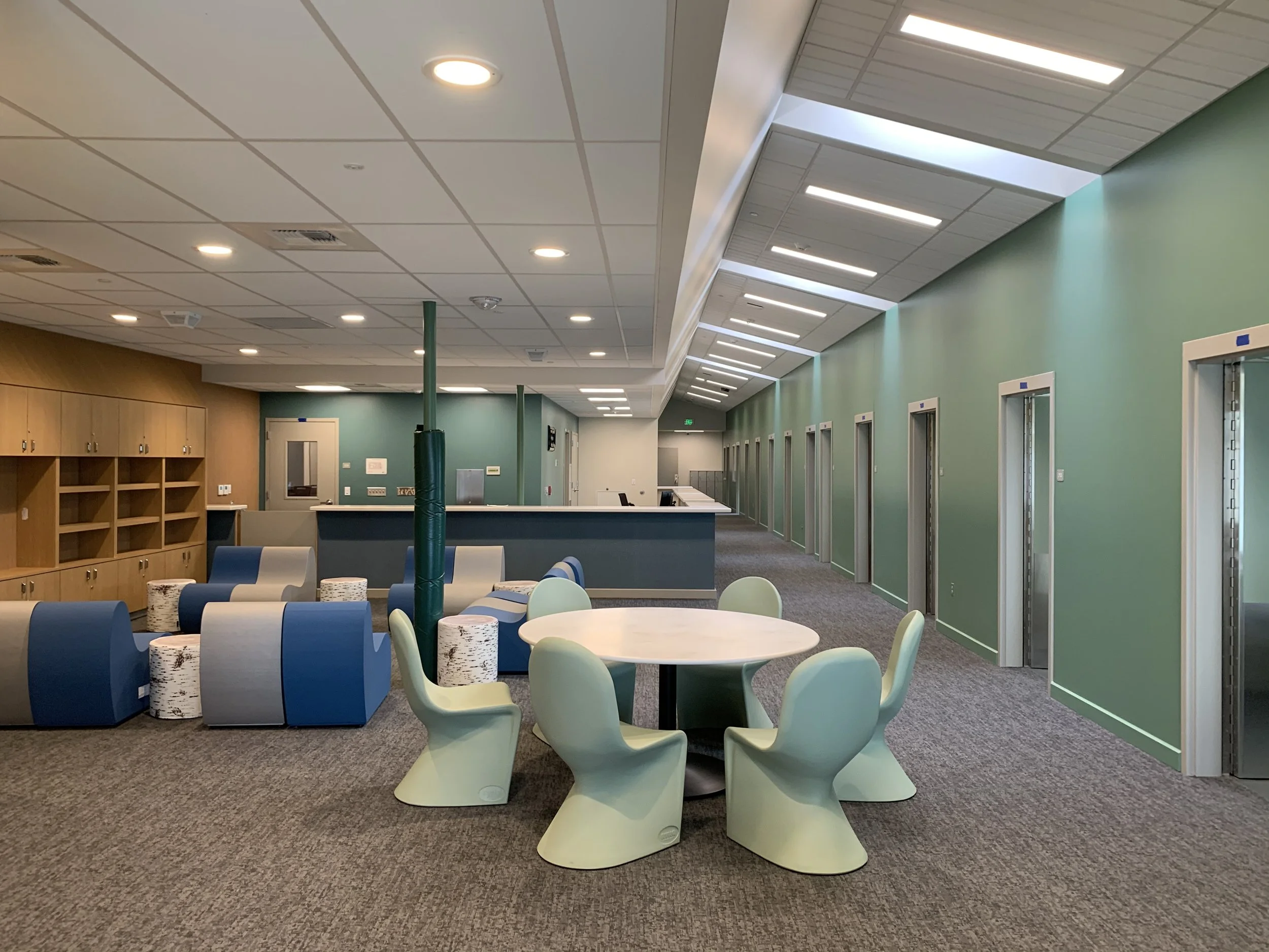 A modern lounge area with circular tables and cream-colored chairs, blue and gray lounge chairs with cylindrical white and black cushions, wooden storage cabinets on the left, a reception counter in the back, and a hallway with multiple elevator doors on the right, all under a ceiling with skylights and recessed lighting.