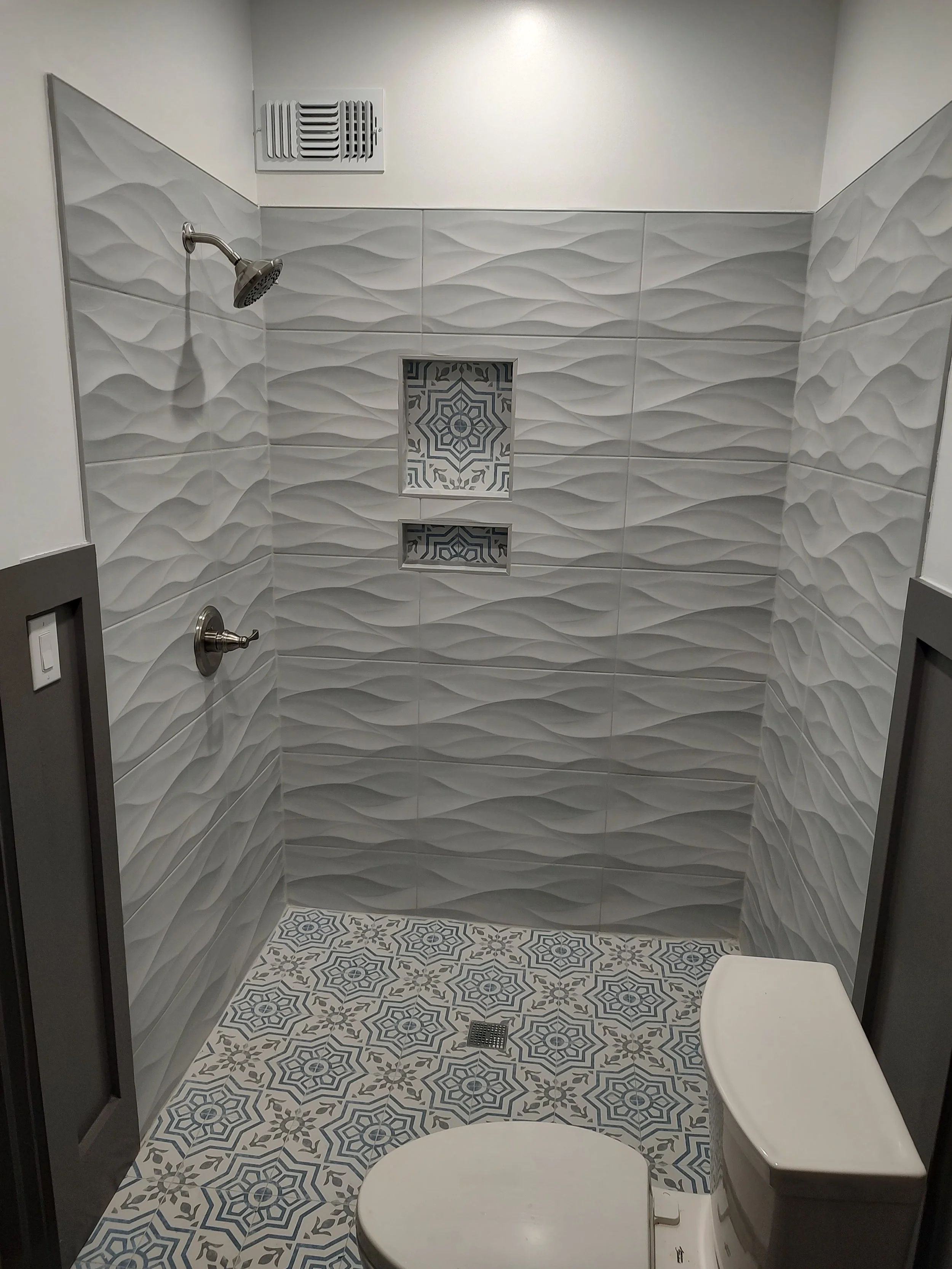 Bathroom shower with textured gray tiles, built-in niche with patterned tiles, showerhead, and a toilet.