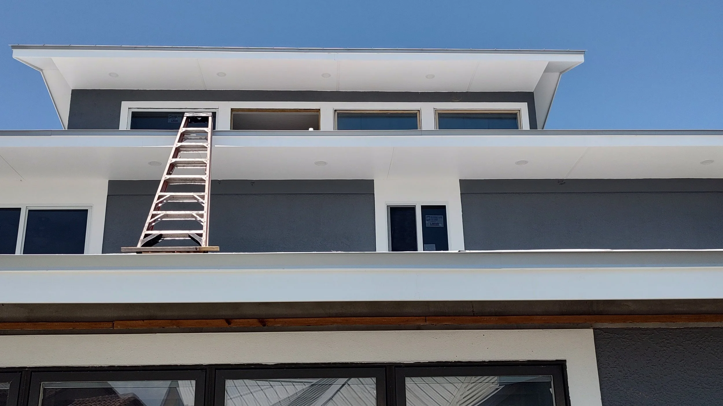 Modern multi-story building under construction with a ladder leaning against the balcony, and construction signs on the windows.