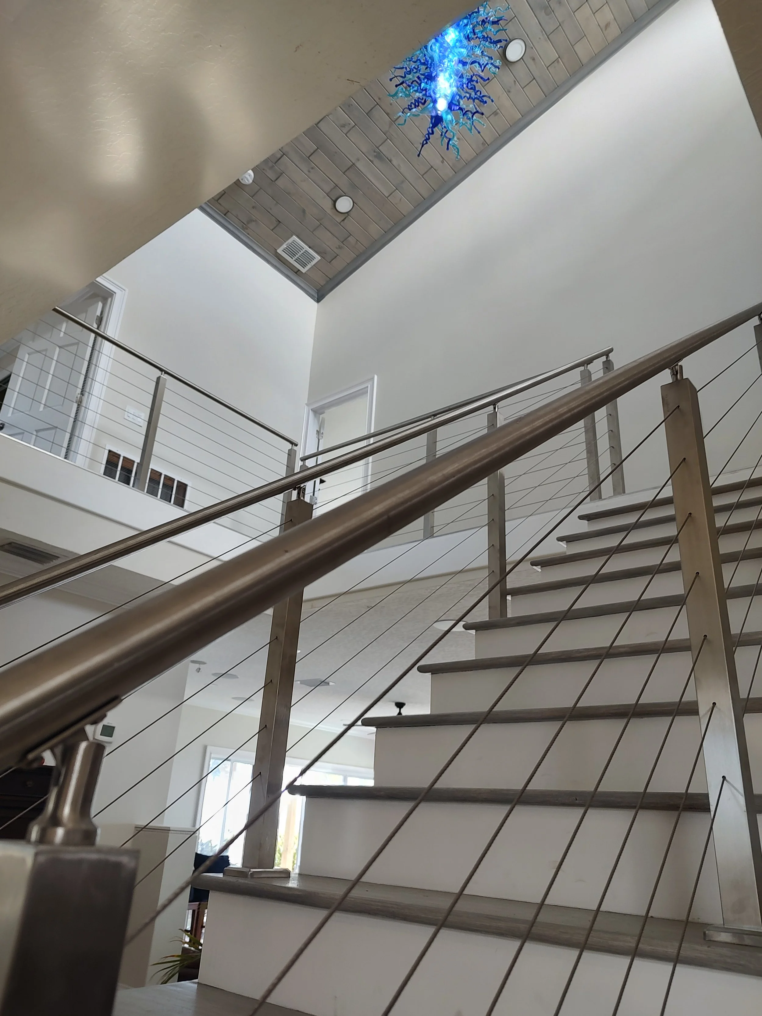 Interior view of a modern staircase with metal railing and wood steps, leading to an upper level with a white door and decorative blue art hanging from the ceiling.