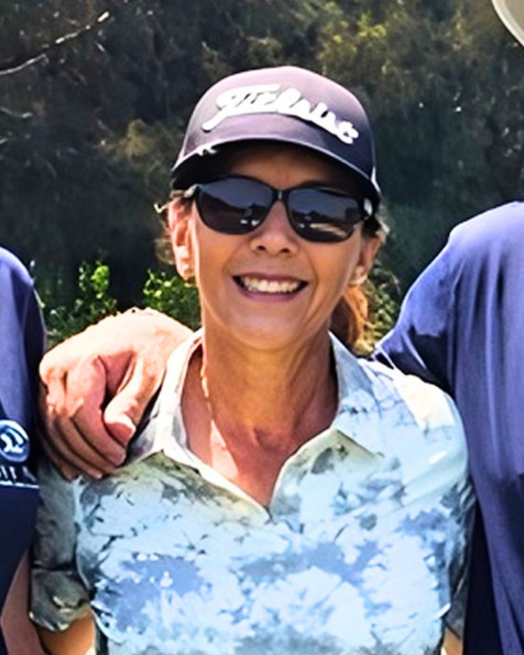 A woman wearing sunglasses, a cap, and a patterned shirt, smiling outdoors with greenery in the background.