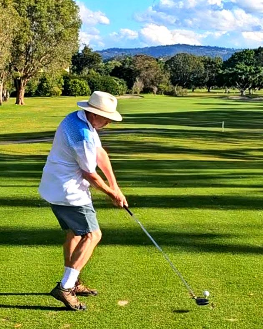 A man in a wide-brimmed hat, white shirt, and shorts preparing to hit a golf ball on a golf course with trees and mountains in the background.