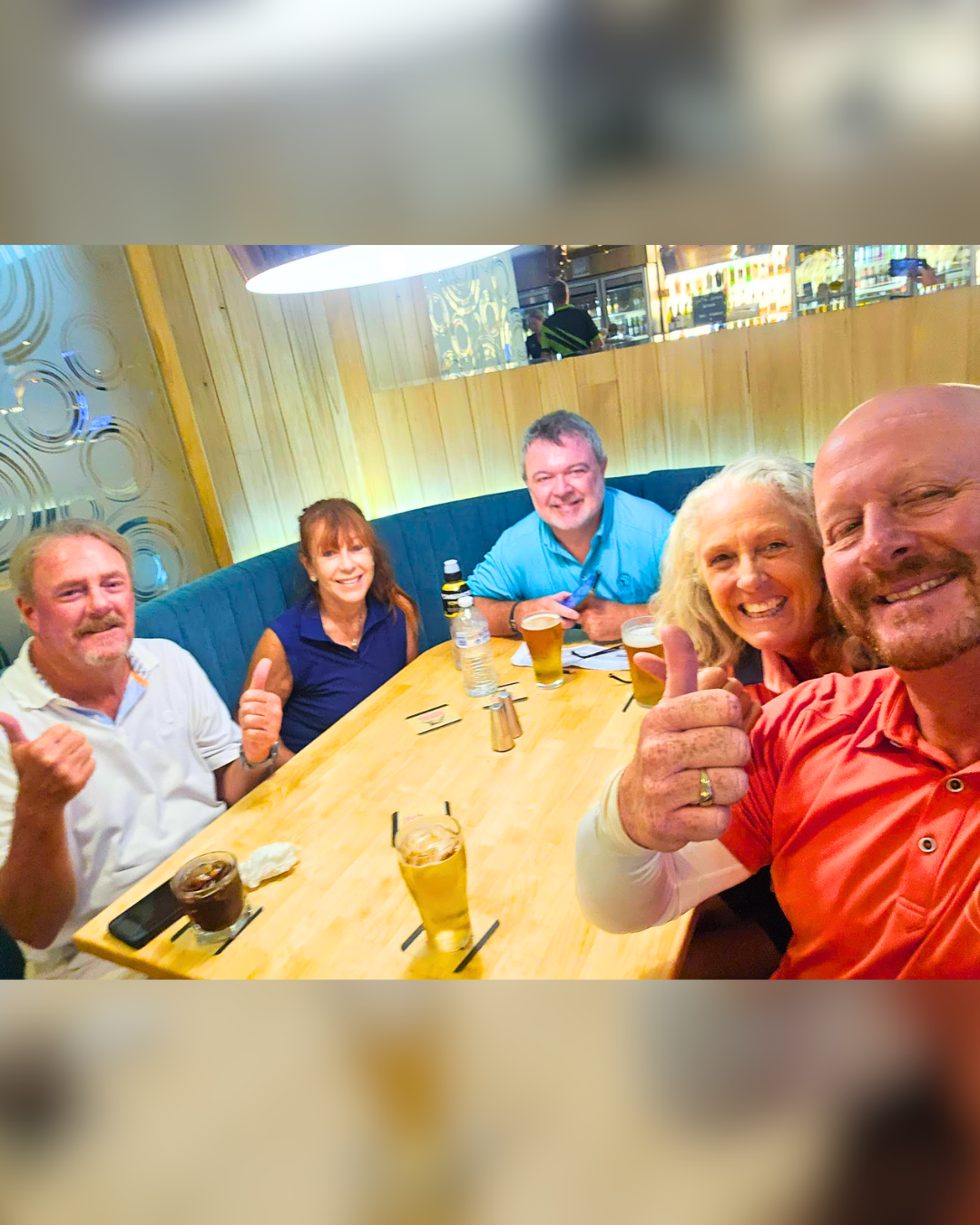 Group of five adults smiling and giving thumbs up at a restaurant table with drinks and snacks.