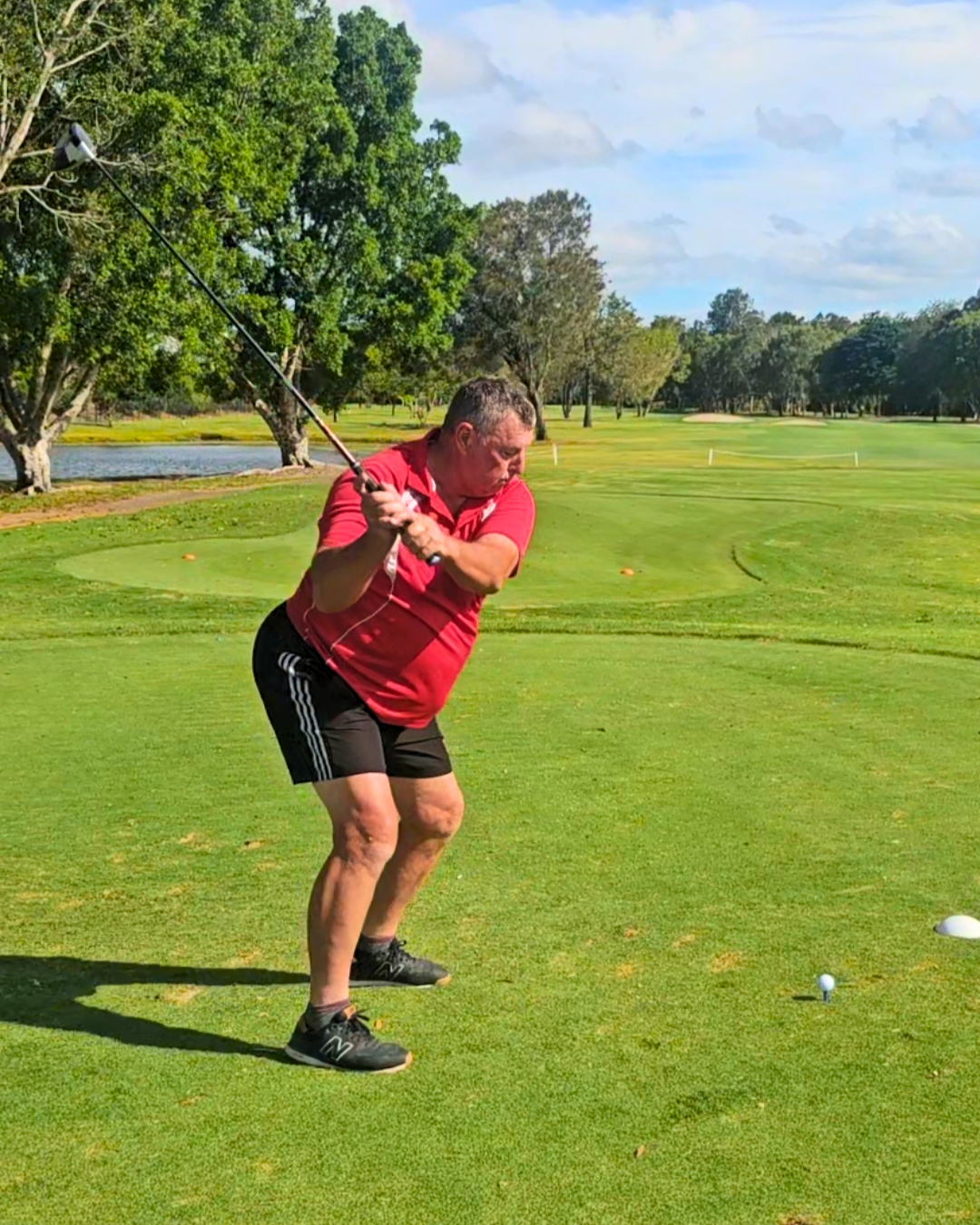 A man wearing a red shirt and black shorts is teeing off on a golf course, preparing to hit a golf ball towards the hole, with trees and a pond in the background.