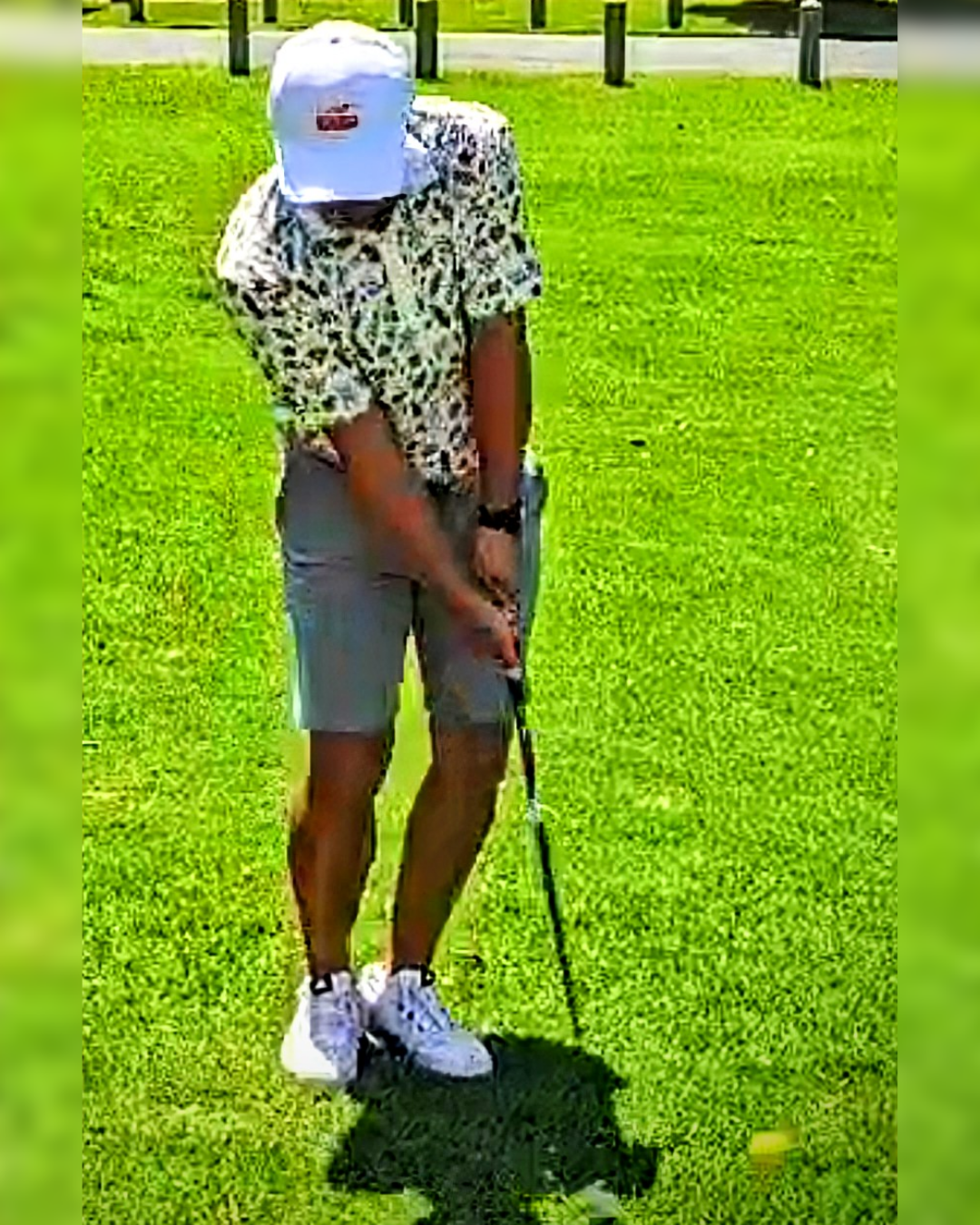 Man playing golf on a green field, wearing a white cap, leopard print shirt, gray shorts, and white shoes, holding a golf club.