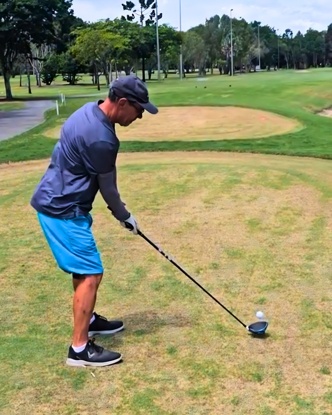 A man in sunglasses, a dark cap, a dark jacket, light blue shorts, and black shoes is preparing to hit a golf ball with a driver club on a golf course, with a sand trap behind him and trees in the distance.