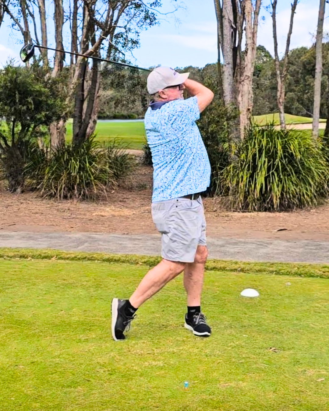A man wearing a light-colored cap, glasses, a blue patterned shirt, gray shorts, and black sneakers is swinging a golf club on a golf course, preparing to hit a golf ball.