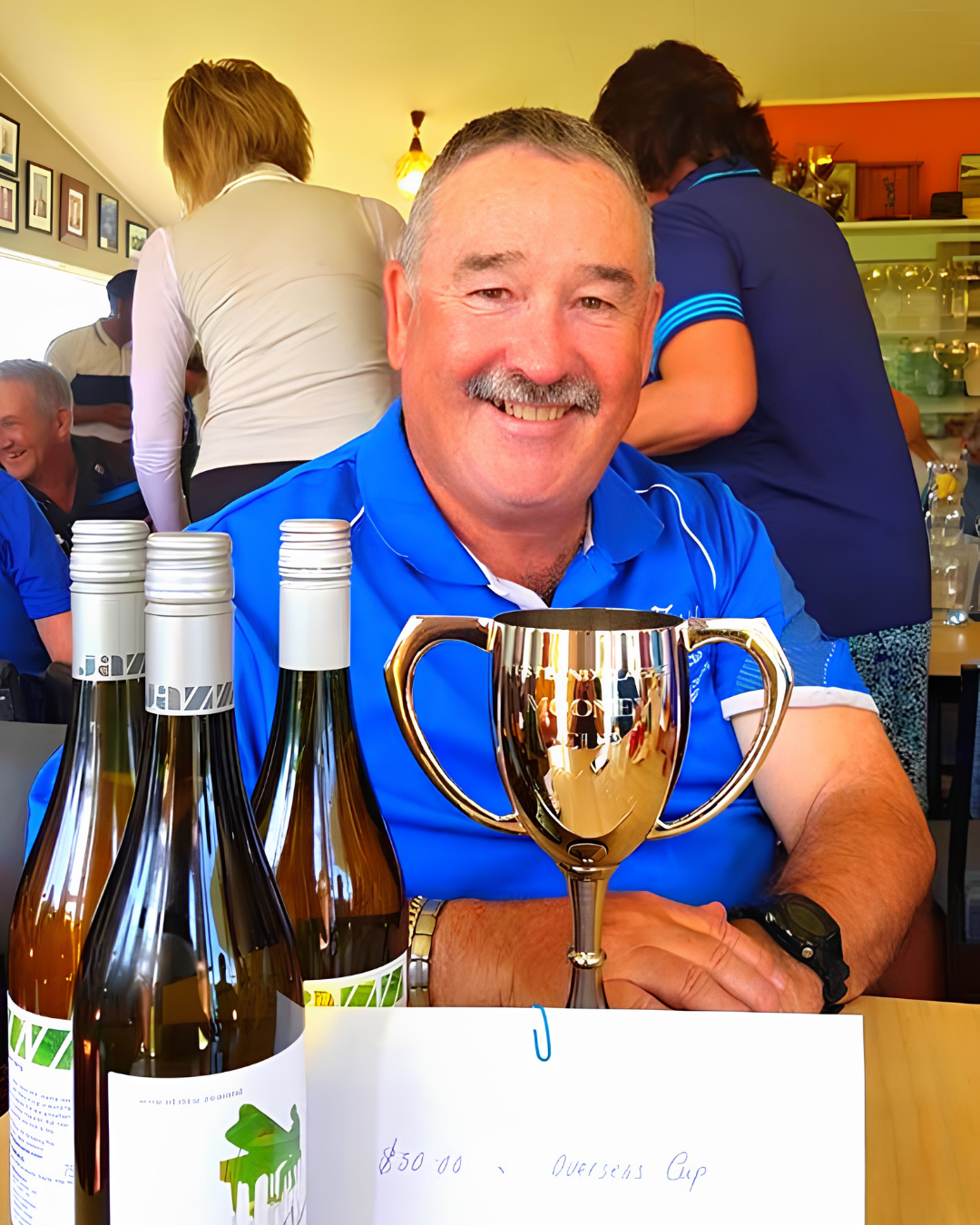 A man in a blue shirt smiling at a table with four bottles of wine, a shiny trophy, and a handwritten note indicating a winning score of 850 and a prize for the winning cup. People are mingling in the background of a lively indoor setting.