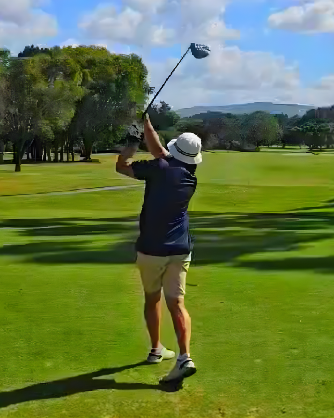 Person playing golf on a sunny day, swinging a golf club on a green golf course with trees and hills in the background.