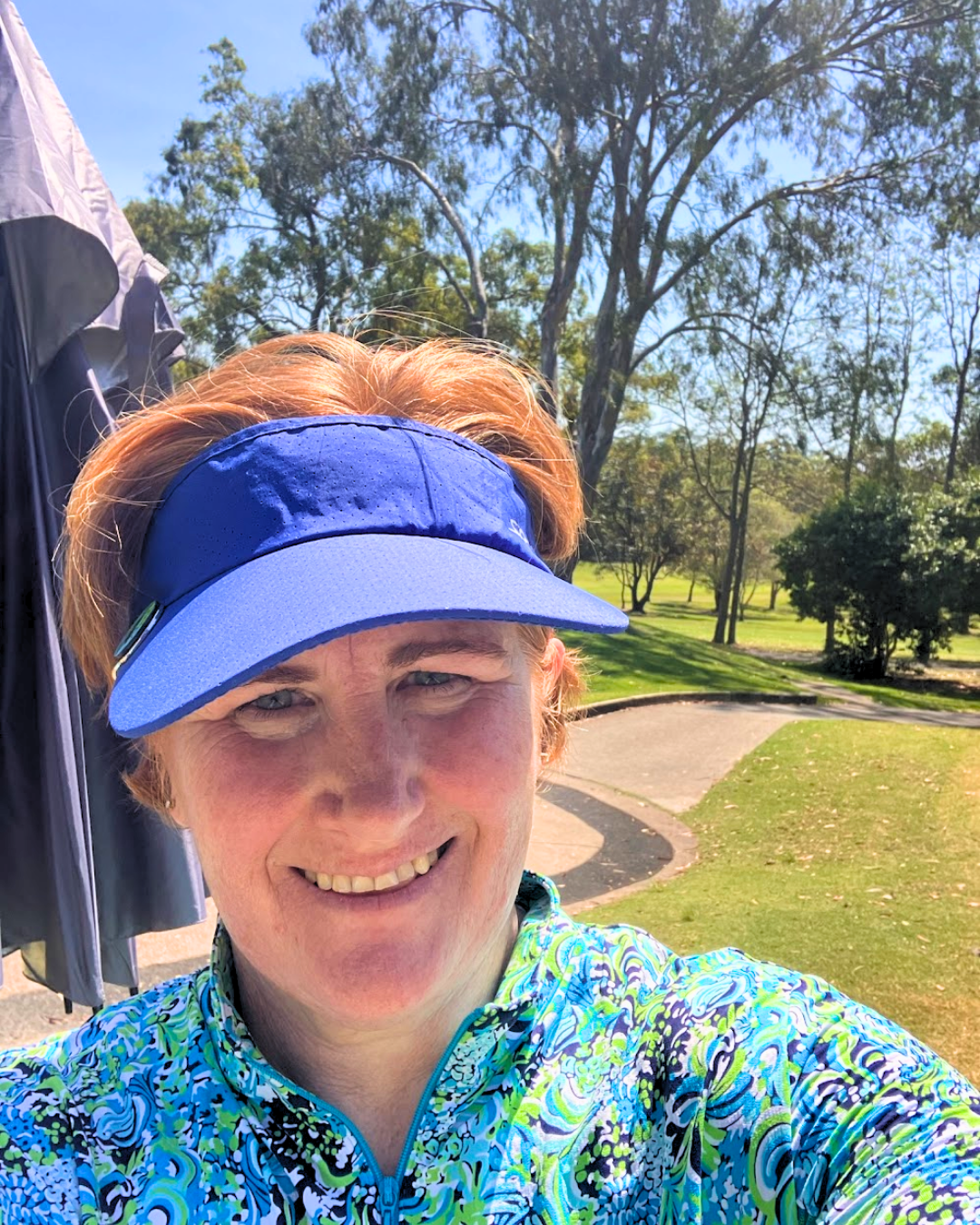 A woman outdoors taking a selfie, wearing a blue visor and a colorful patterned athletic jacket, with trees and a walking path in the background on a sunny day.