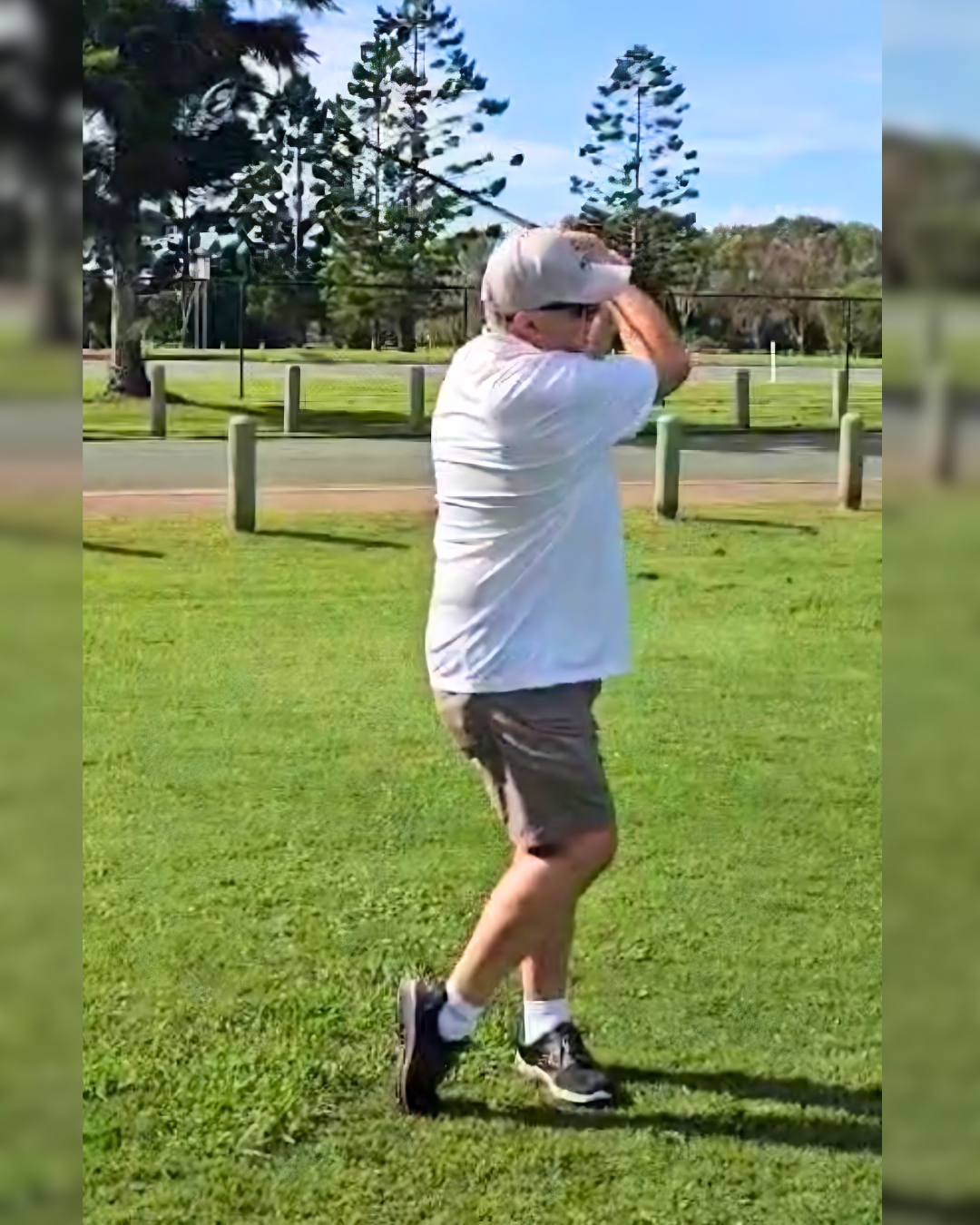 A man standing on grass in a park, holding a baseball bat and preparing to swing, wearing a white cap, glasses, white t-shirt, khaki shorts, and sneakers during daytime.