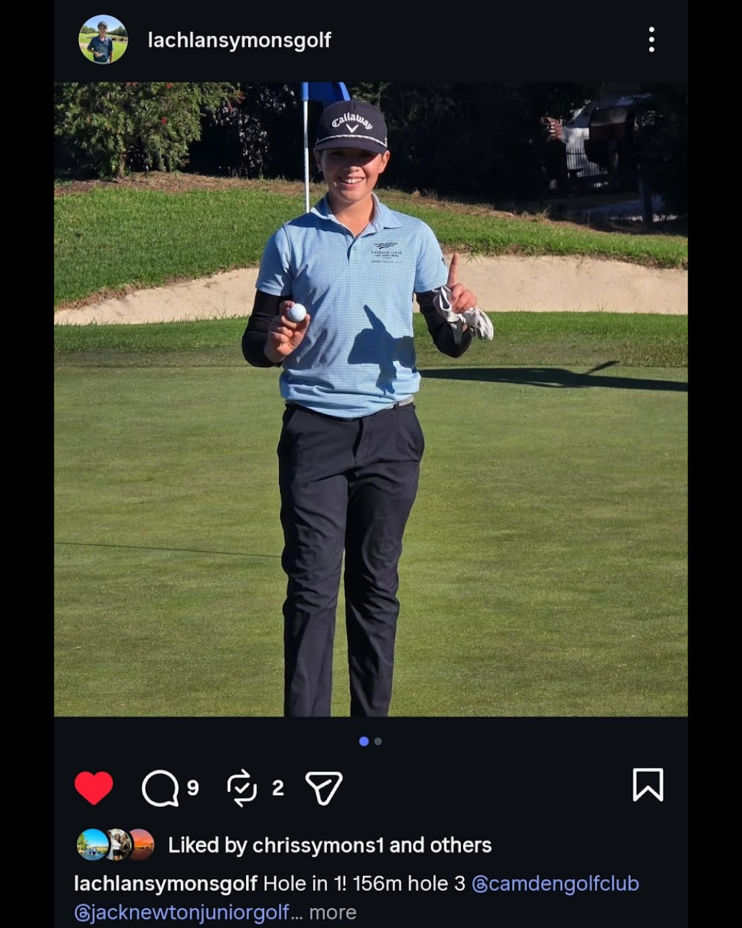 A young person standing on a golf course holding a golf ball and pointing upward with their right index finger, smiling, dressed in golf attire with a Callaway cap, with a golf flag in the background.