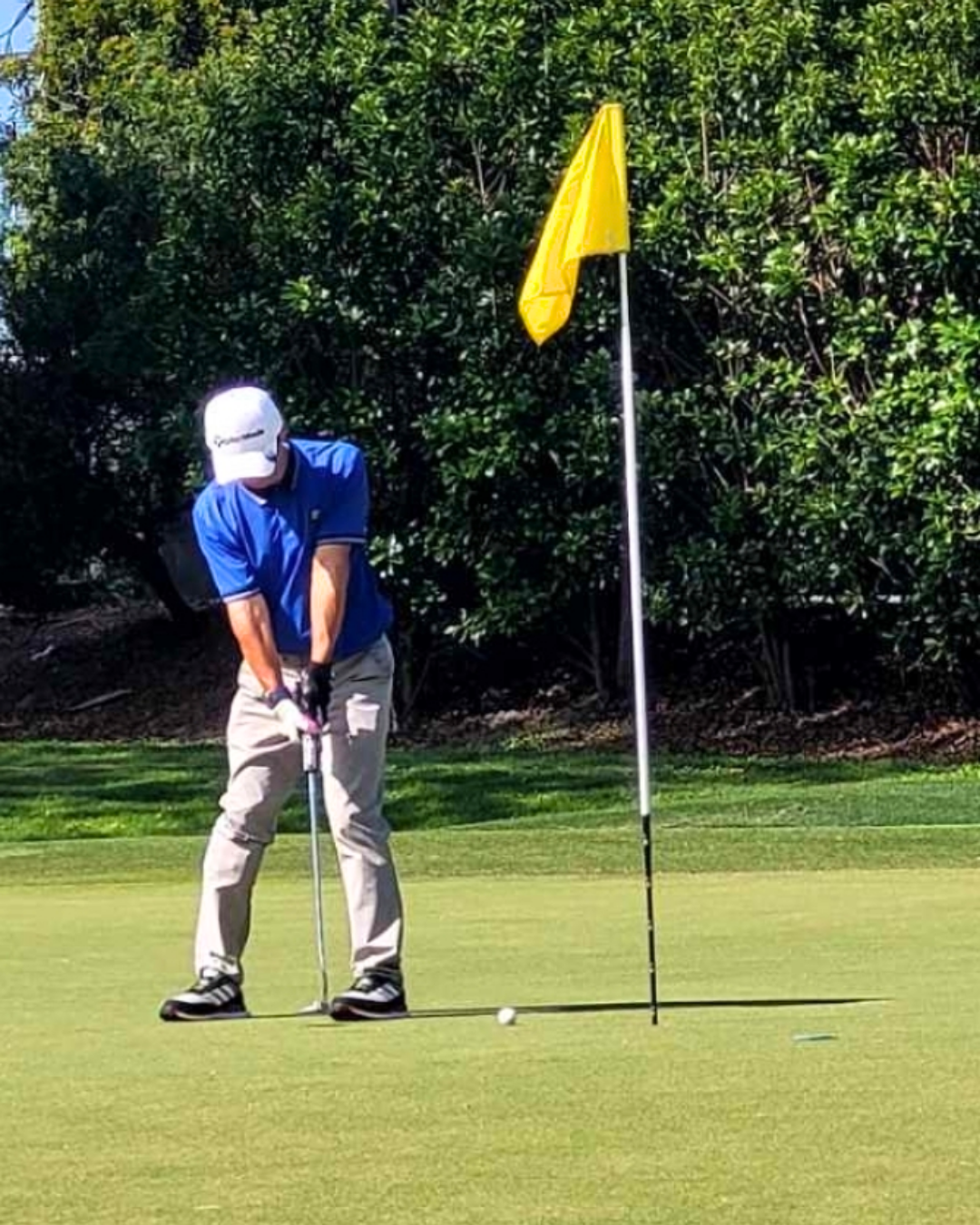 A person in a blue shirt and khaki pants preparing to putt on a golf green near the hole with a yellow flag, with trees in the background.