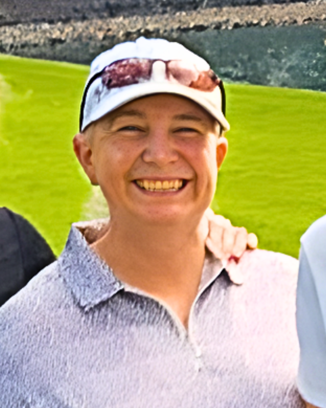 Smile of a man wearing a beige cap with sunglasses on top and a light-colored collared shirt, standing outdoors on a sunny day with a green field and a stone wall in the background.