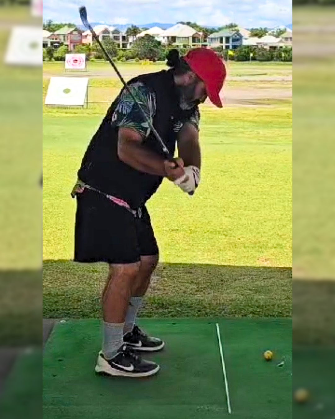 A man wearing a red cap, black shorts, and a black shirt with a floral pattern is preparing to hit a golf ball on a green course. He is holding a golf club and positioned next to a yellow golf ball.