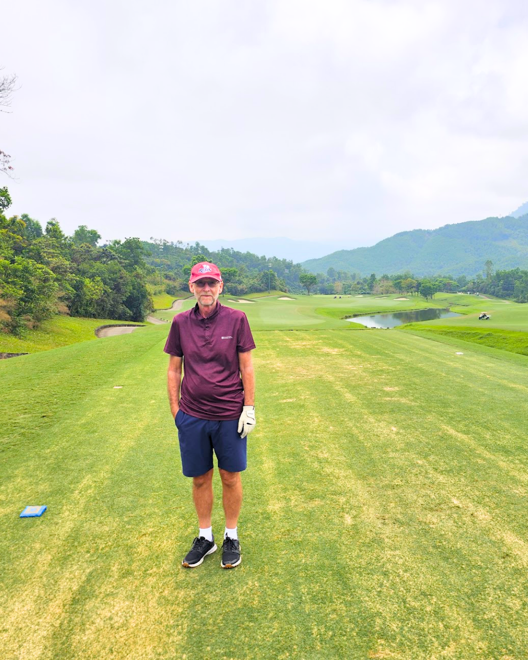 A man standing on a golf course with mountains and trees in the background, wearing a maroon shirt, navy shorts, and a red cap.