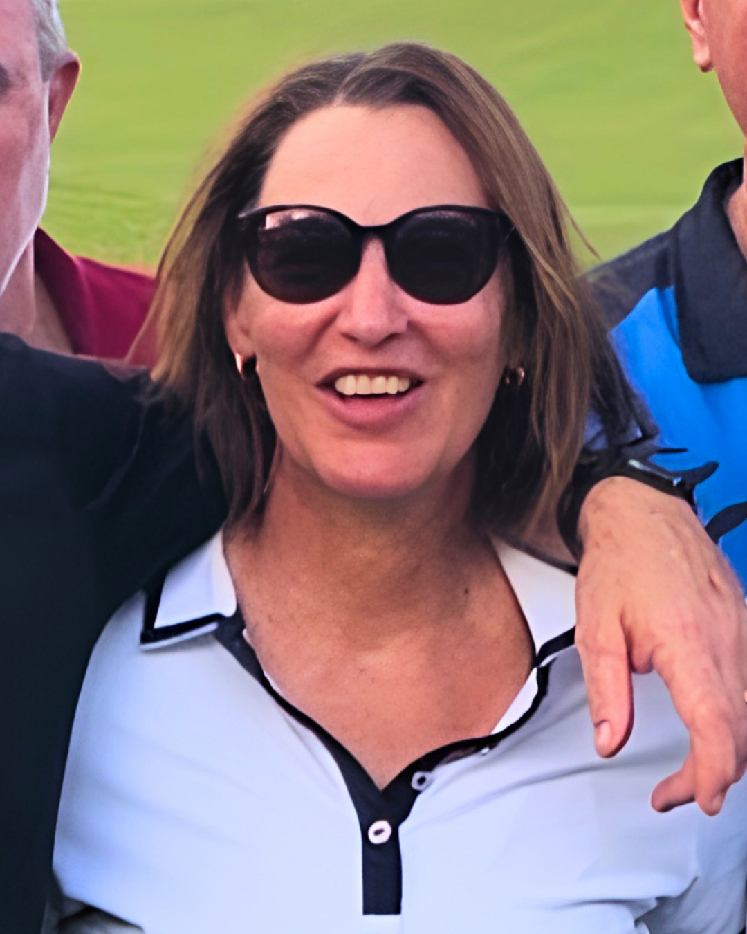 Close-up of a woman with brown hair wearing sunglasses and a white and black collared shirt, smiling, with two people partially visible beside her.