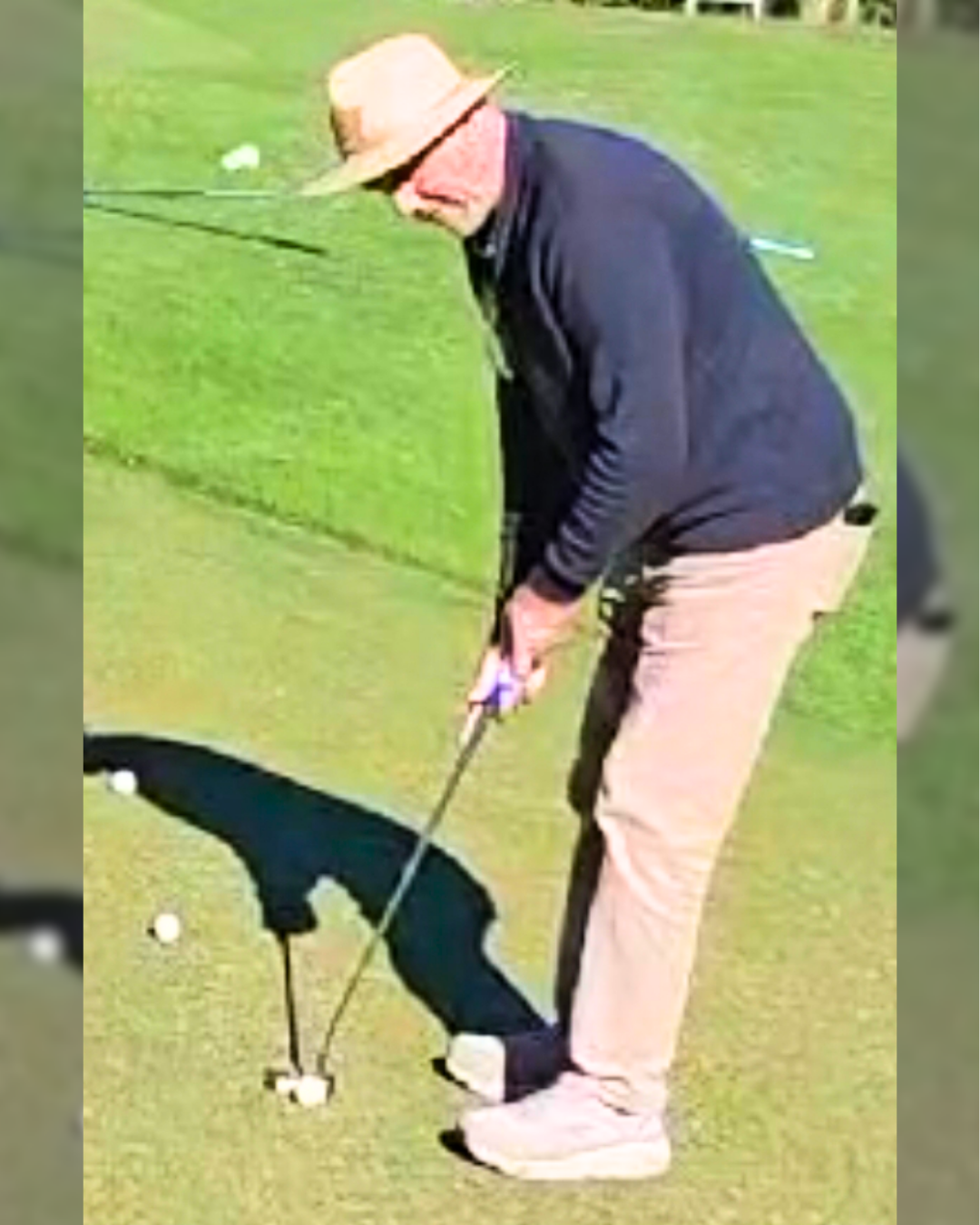 Person wearing a hat, black jacket, and beige pants, putting a golf ball on a tee on a putting green, with golf balls scattered around.