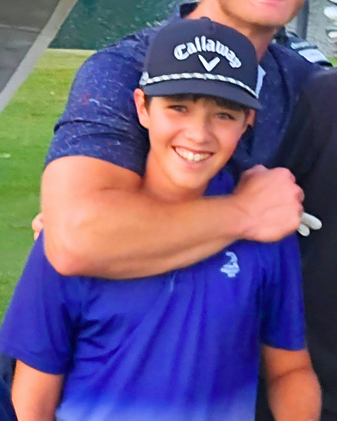 A young man smiling and giving a friendly hug to someone wearing a Callaway cap and a blue shirt outdoors on a grassy field.
