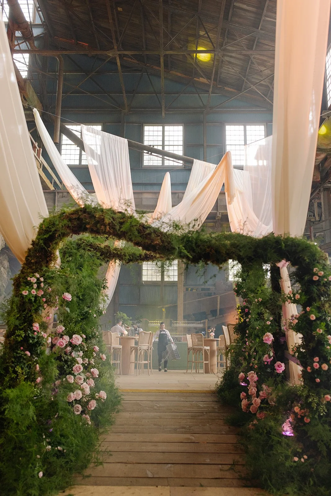 A decorated indoor space with a floral archway leading to a seating area with tables and chairs, set inside an industrial-style building with large windows and draped fabric decorations hanging from the ceiling.