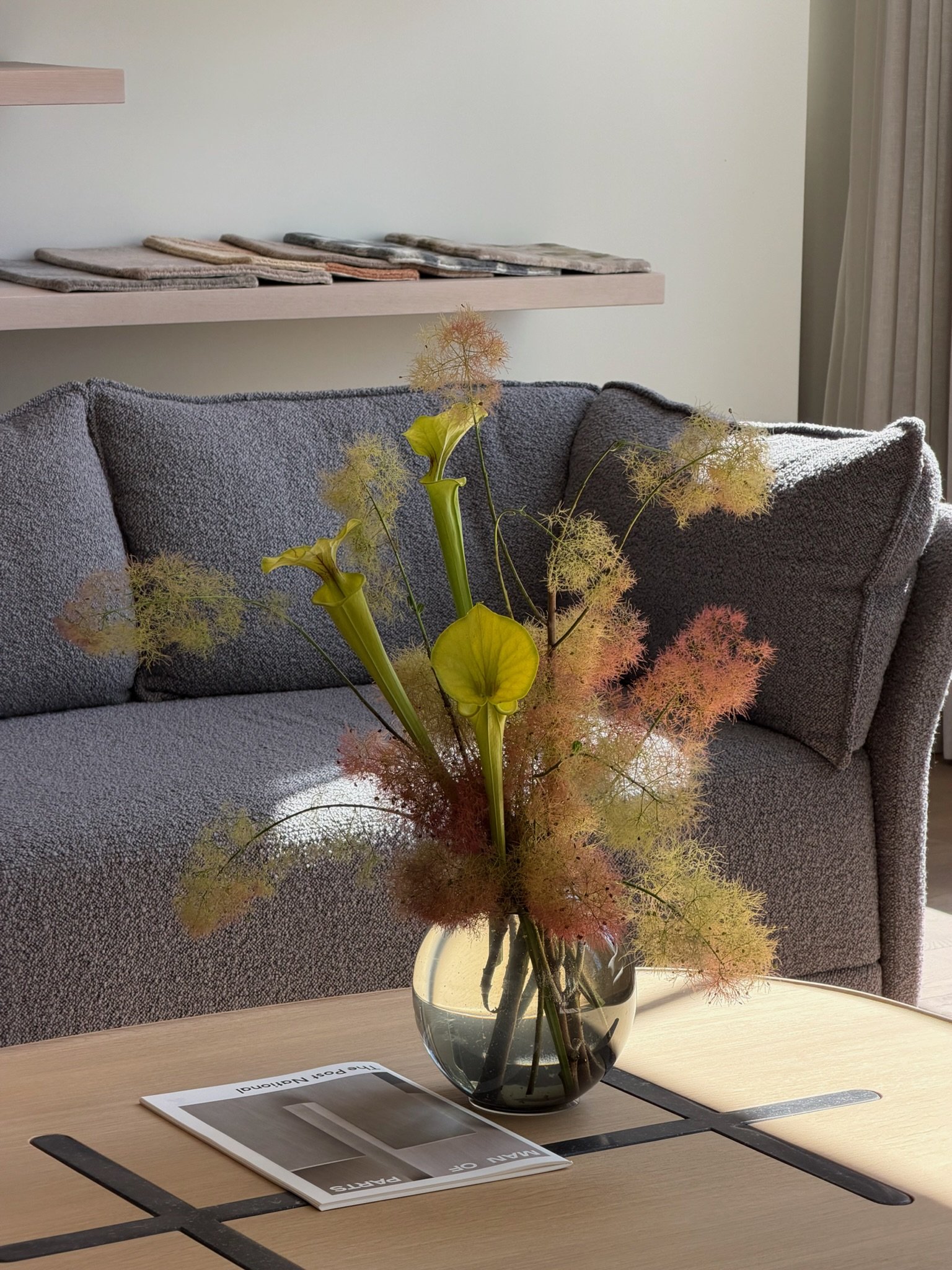 A clear glass vase on a wooden table holds a bouquet of colorful, fluffy flowers with light green and yellow leaves. Behind the table, there is a gray fabric sofa, and in the background, a white wall with a wooden shelf holding various fabric samples