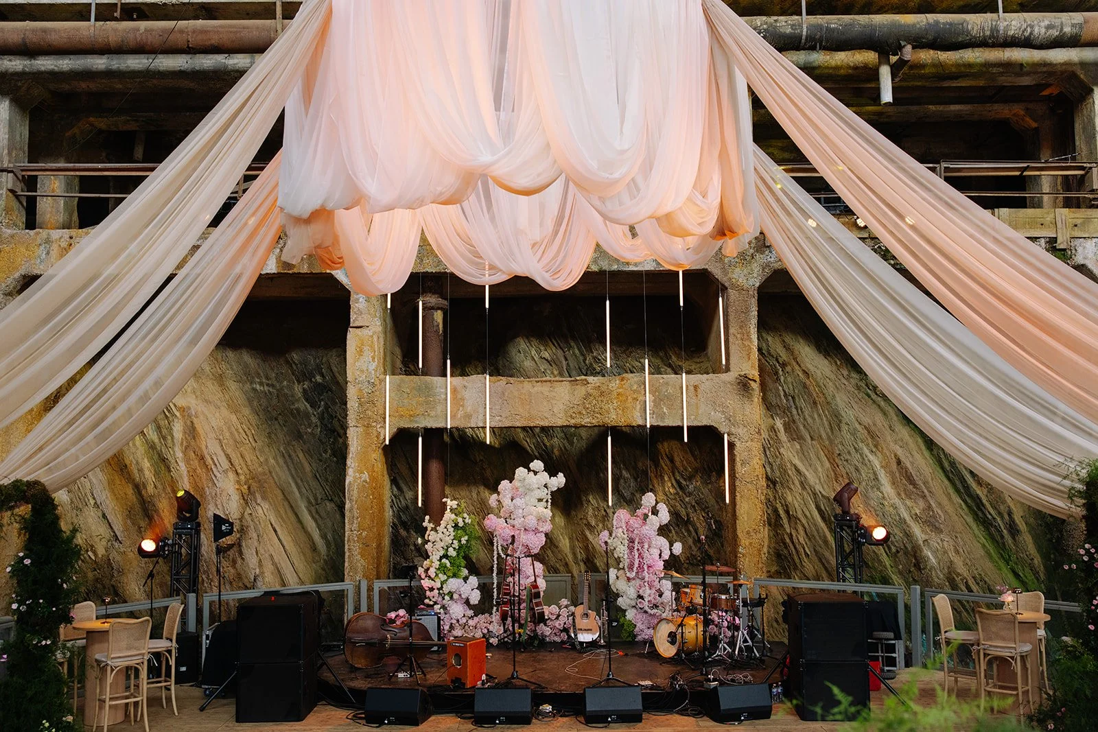 Decorative stage setup with pink and white drapes hanging from rock wall, floral arrangements, musical instruments, and event lighting in an outdoor rustic setting.