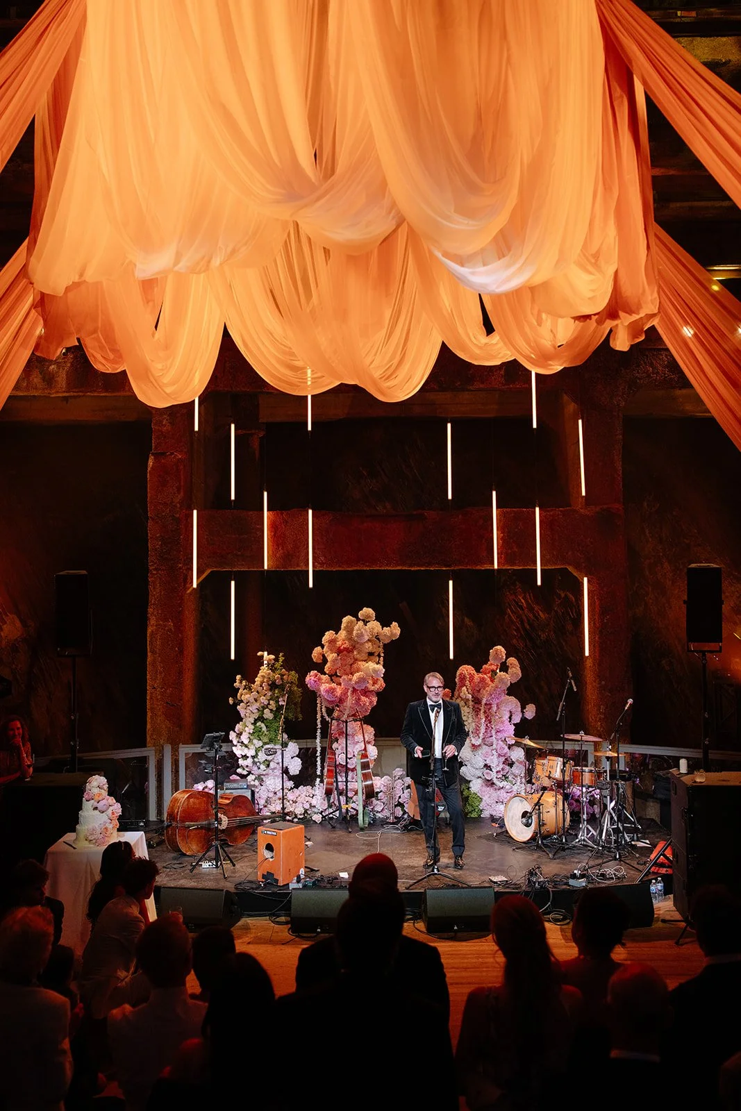 A man in a tuxedo and glasses performing on stage at a wedding or event venue, with floral arrangements and musical instruments, under large peach-colored drapery and vertical light fixtures.