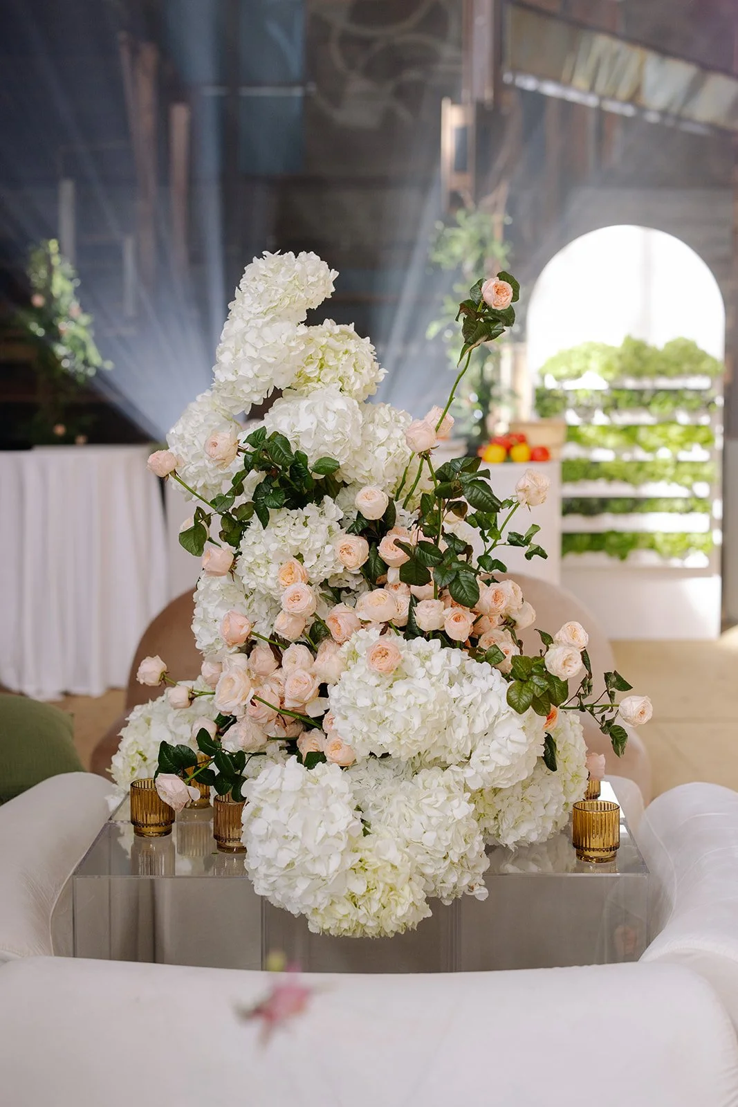 A floral centerpiece made of white hydrangeas and pale pink roses placed on a glass table in a bright room with natural light.