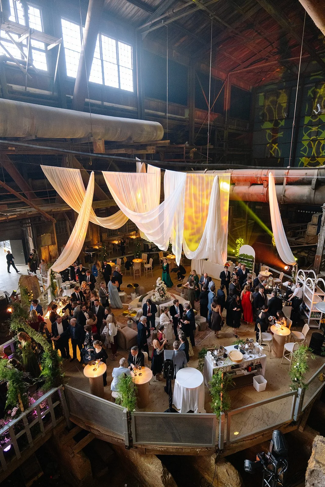 Indoor event in a large industrial space, decorated with flowing white fabric, greenery, and warm lighting, with many people dressed formally, standing and socializing around tables and a bar area.