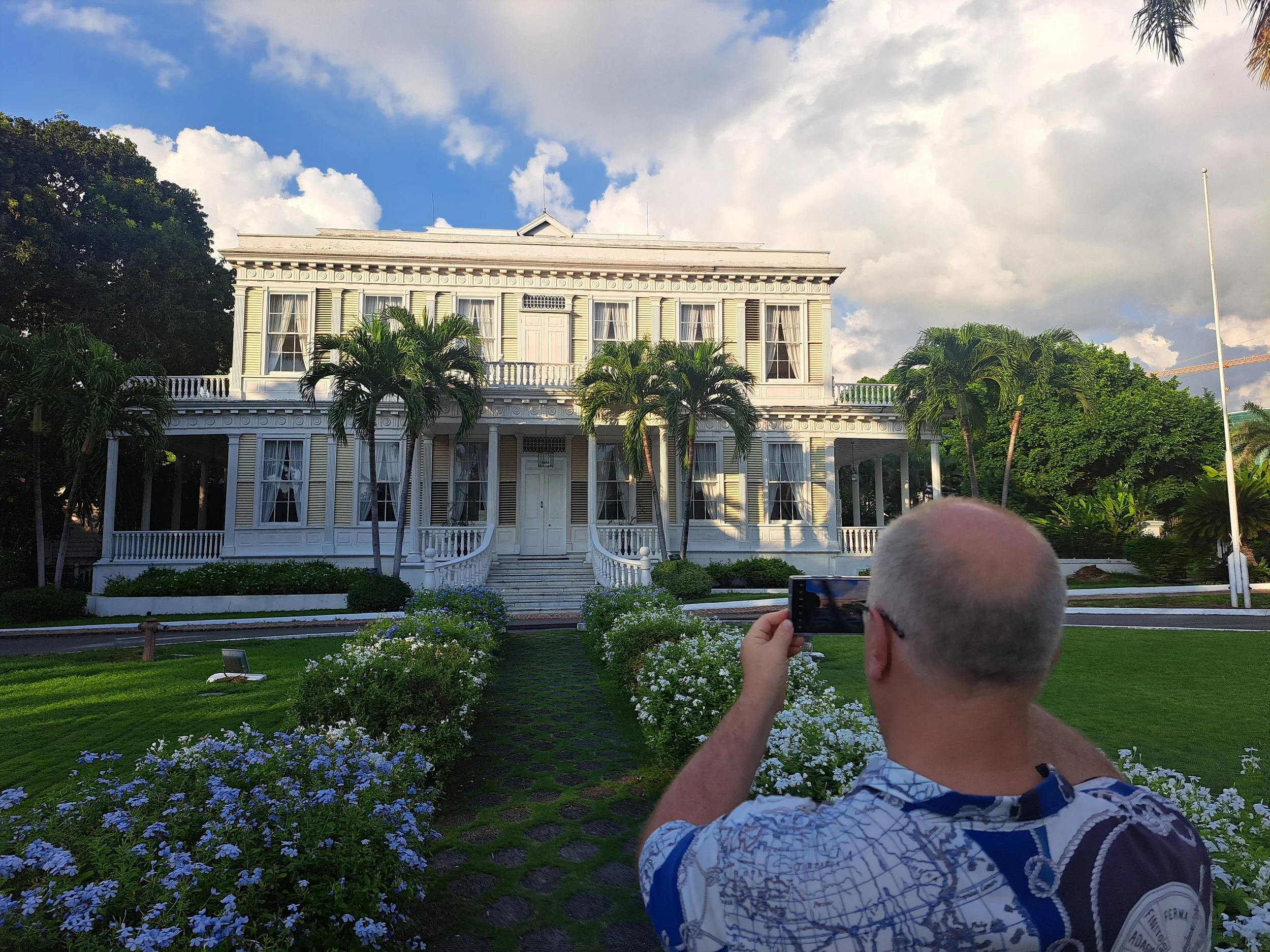 A man with gray hair taking a photo of a large white mansion with palm trees and a manicured garden under a partly cloudy sky. Kgn jamaica