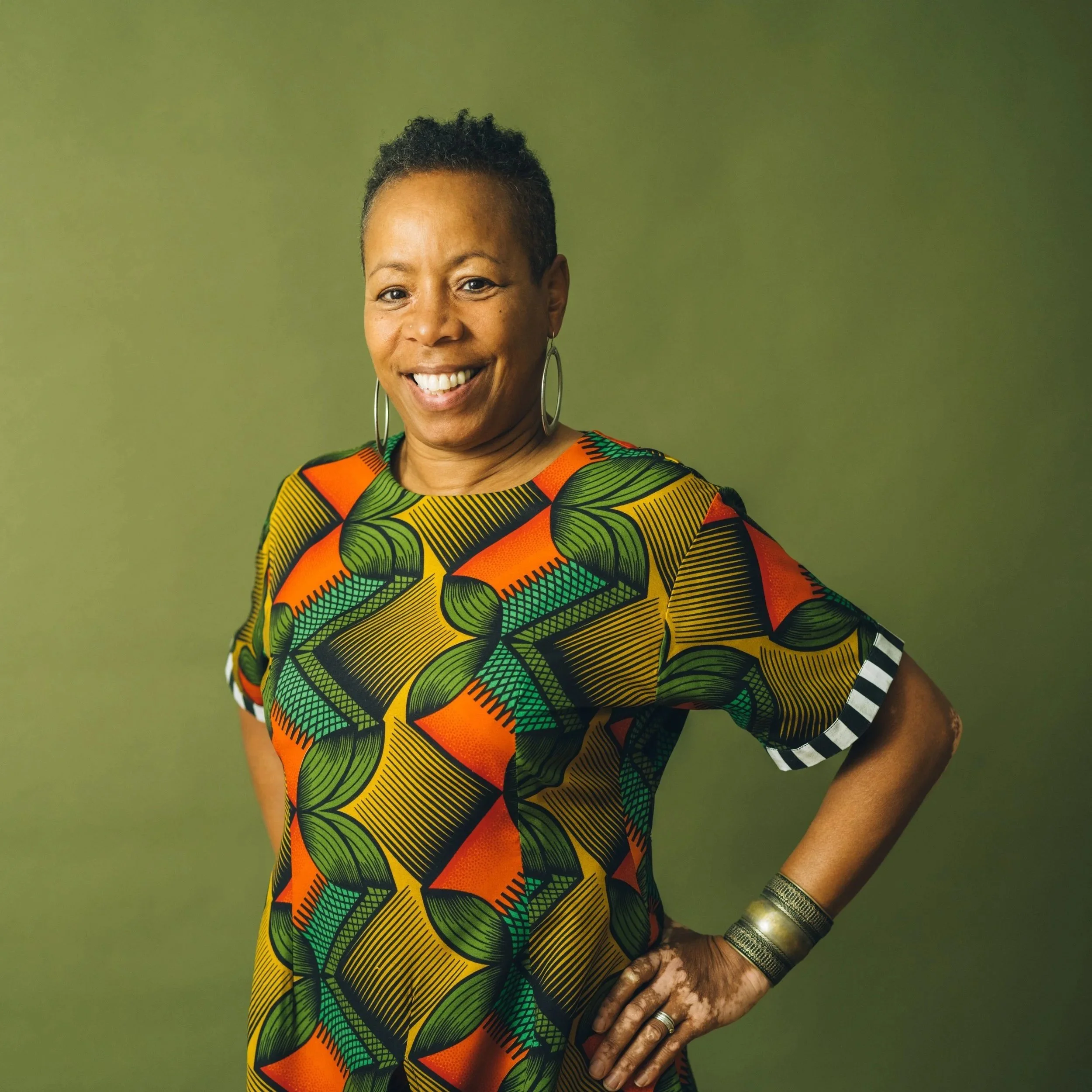 Portrait of a woman with short black hair, smiling, wearing large earrings, a colorful dress with geometric patterns in green, yellow, orange, and black, against a plain green background.