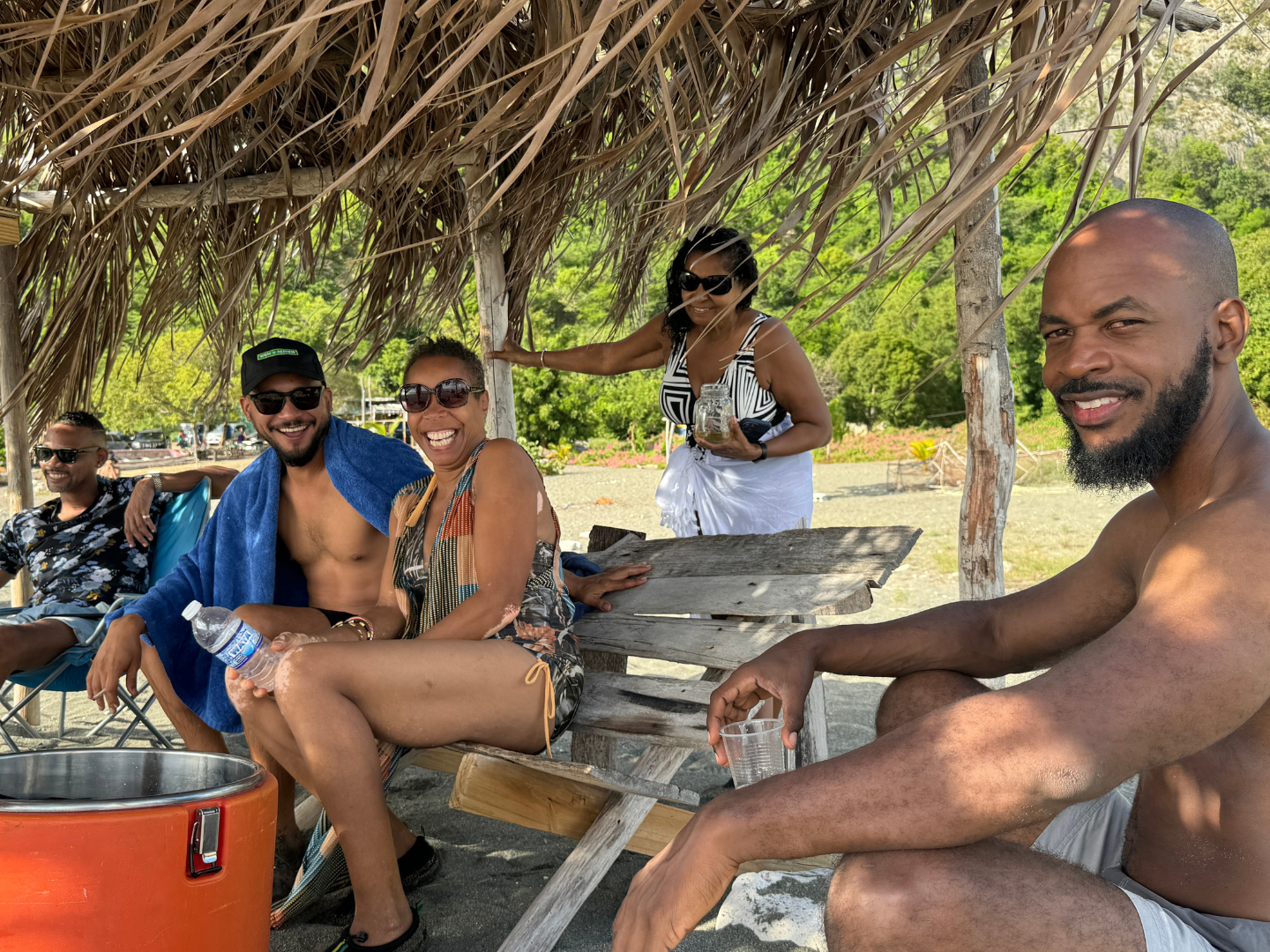 Group of friends relaxing at Bob Marley beach in Bull bay Kingston, St Andrew Jamaica under a thatched roof, smiling and enjoying drinks on a sunny day.