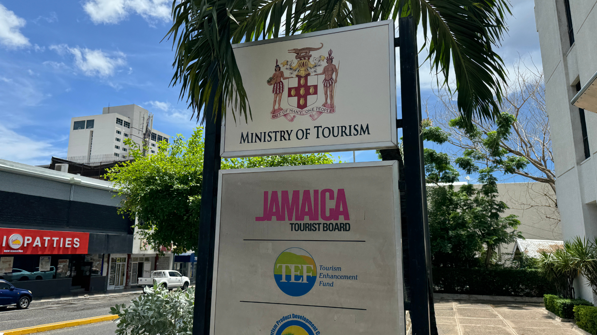 Signboard of the Ministry of Tourism and Jamaica Tourist Board with a crest at the top, set outdoors on a sunny day with trees and buildings in the background.