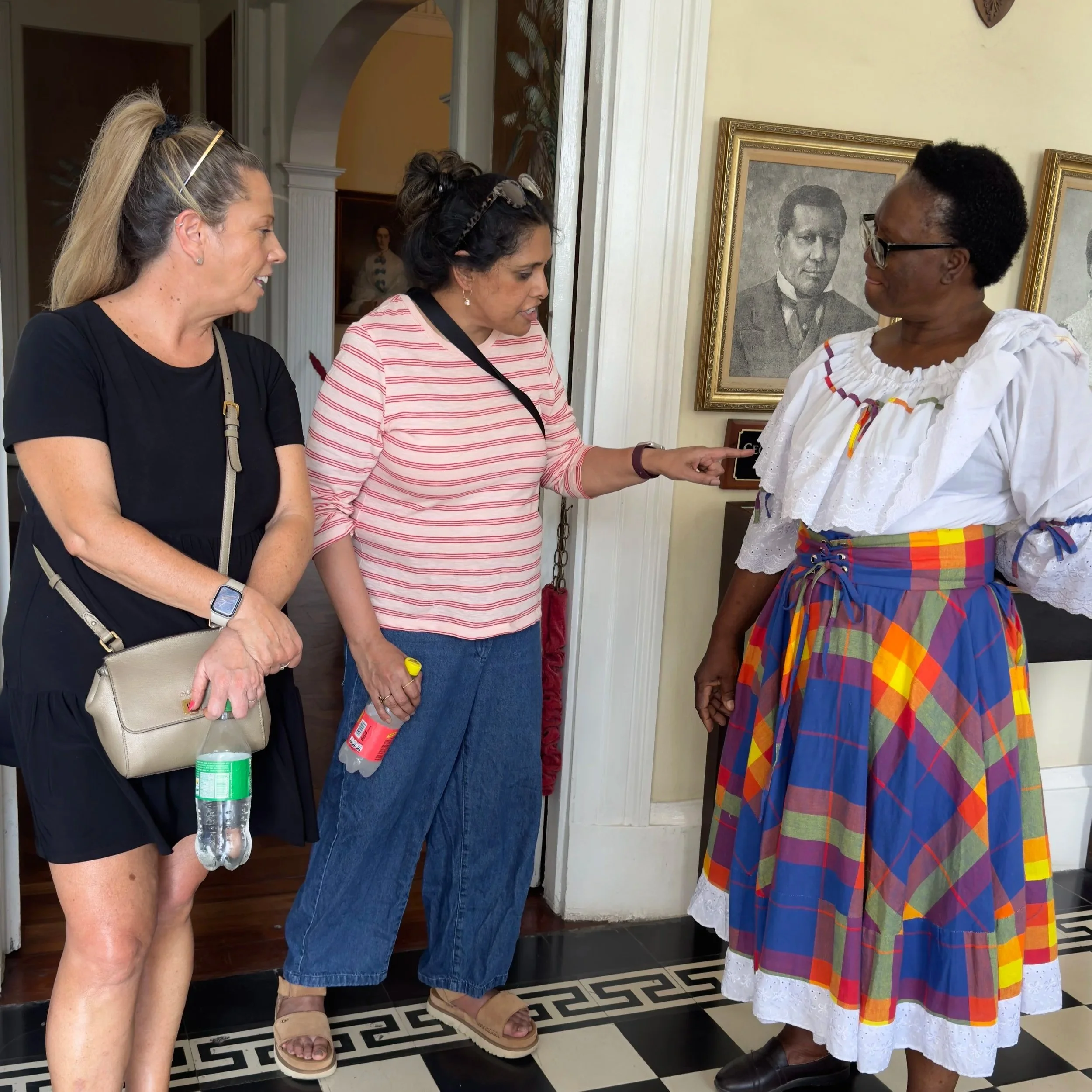 Three women having a conversation indoors, with framed portraits on the wall behind them.