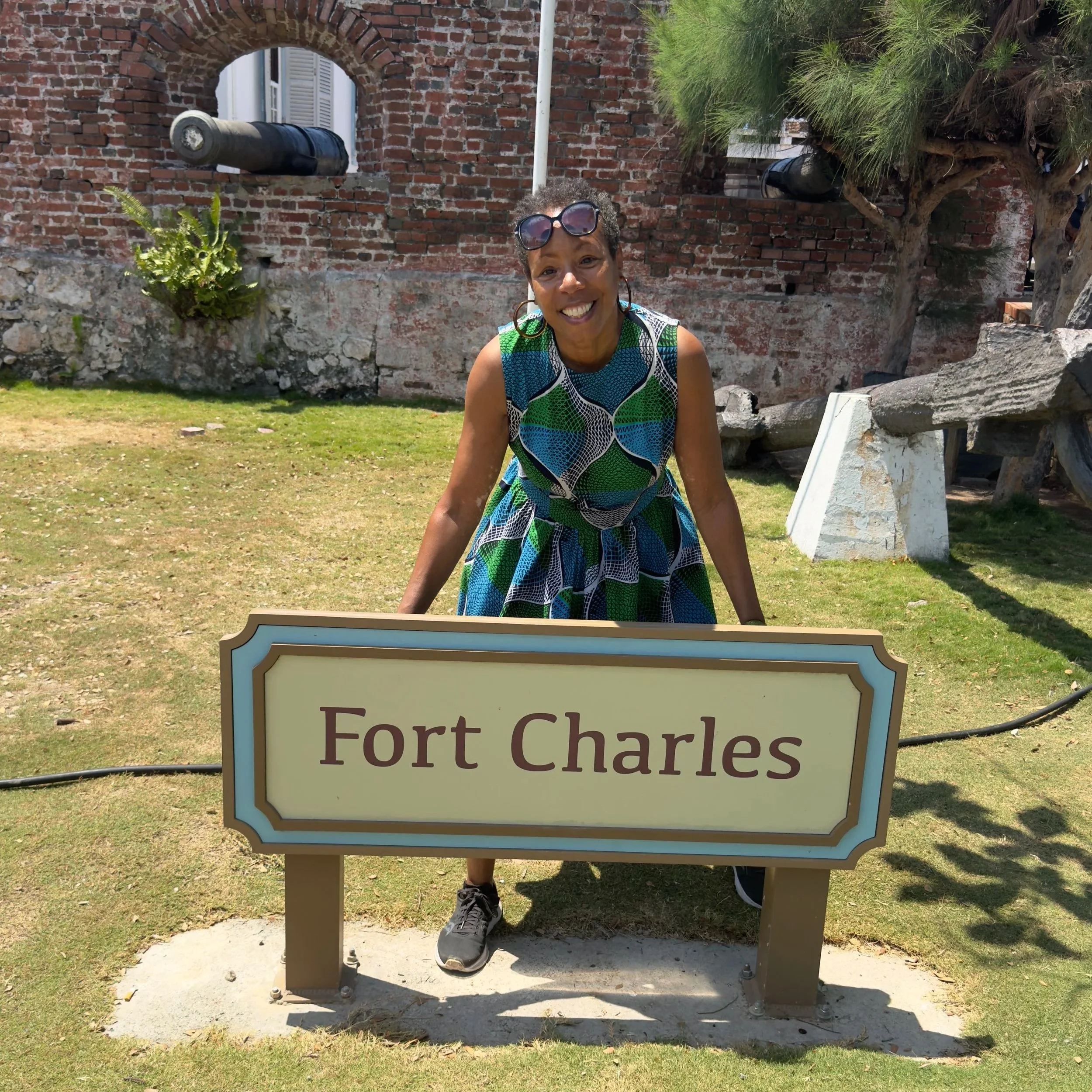 A smiling woman in a colorful dress stands behind a sign that reads 'Fort Charles' on a grassy area near a weathered brick wall and a tree. While on tour in Fort Charles Port Royal Jamaica with Onestop cultural tours Jamaica.