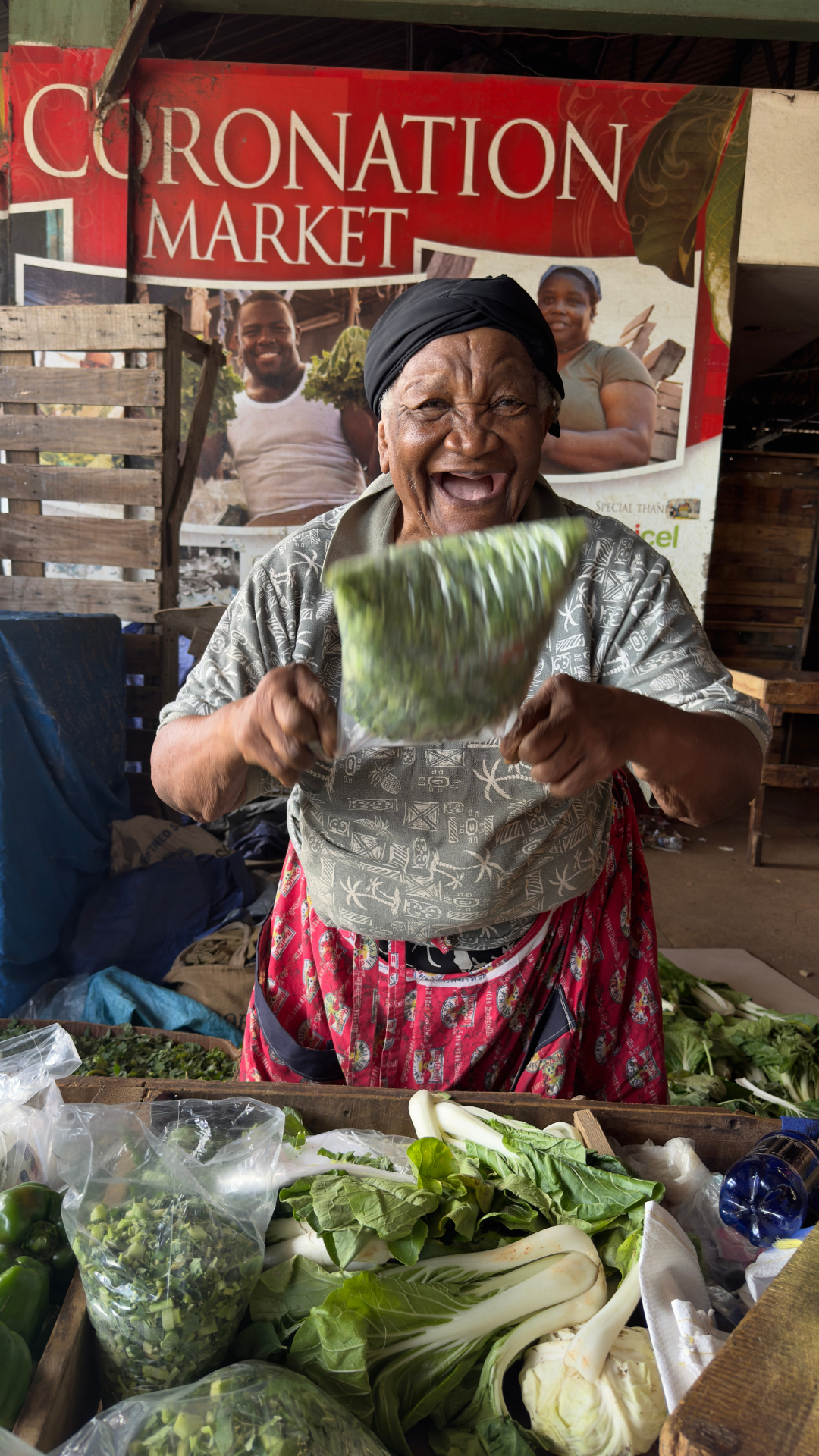 An elderly woman joyfully selling fresh vegetables at Kingstons downtown market with a 'Coronation Market' sign in the background.