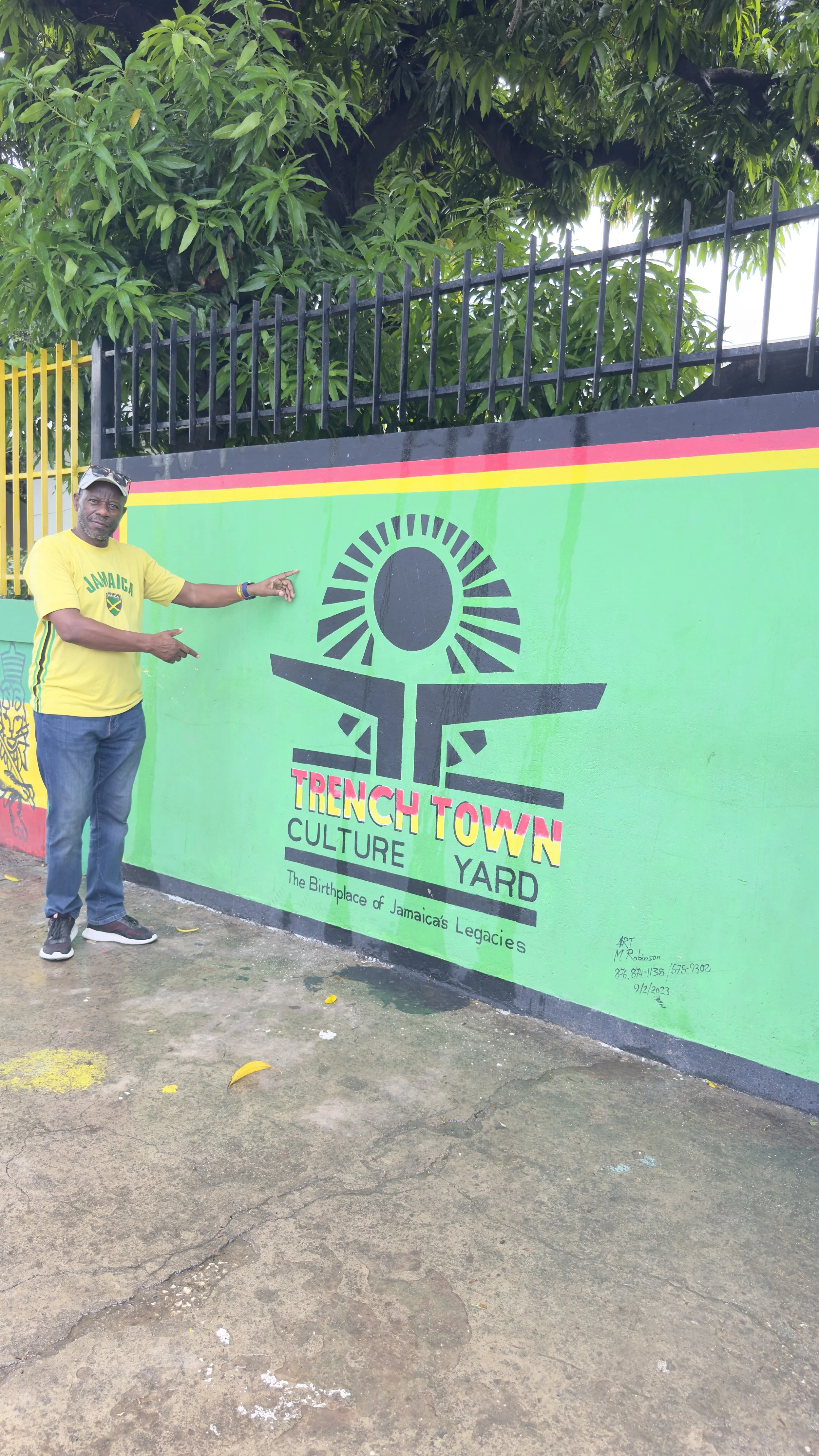 A man in a yellow Jamaica shirt pointing to a colorful mural promoting Trench Town Culture Yard in Jamaica.