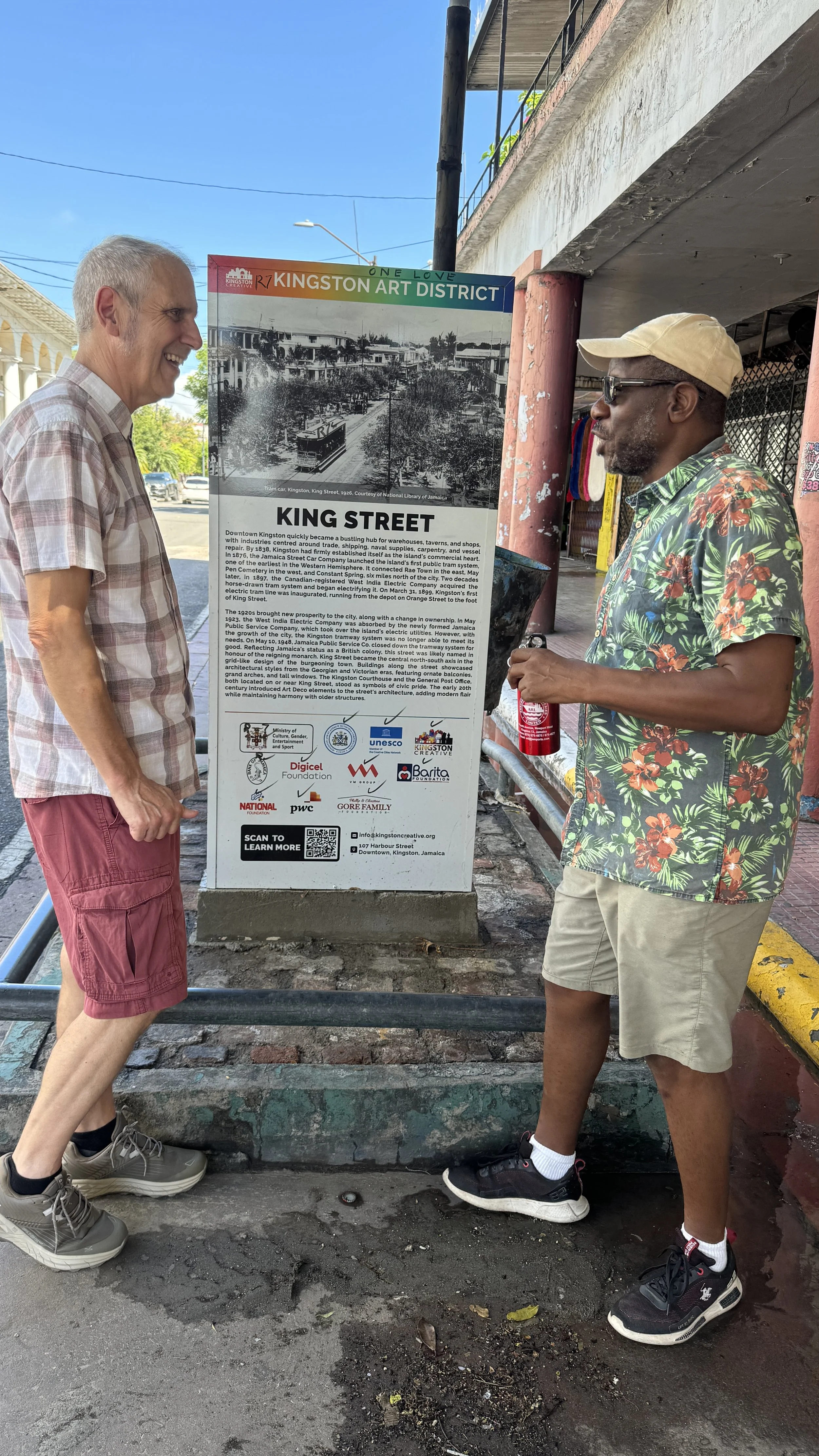 Two men standing on a sidewalk beside an informational sign about Kingston's King Street. The man on the left is wearing a plaid shirt and red shorts, while the man on the right is wearing a floral shirt, beige shorts, a cap, and sunglasses. They are