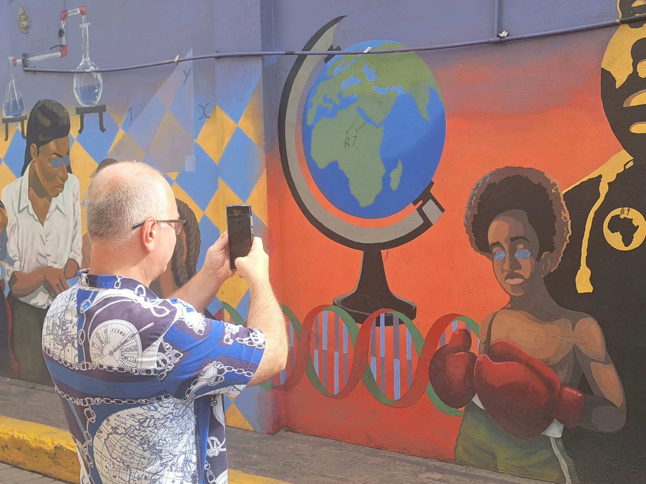 Man taking a photo of a colorful mural featuring a woman with boxing gloves, a globe, DNA strands, and children. while on tour with onestop cultural tours Jamaica in downtown Kingston Jamaica